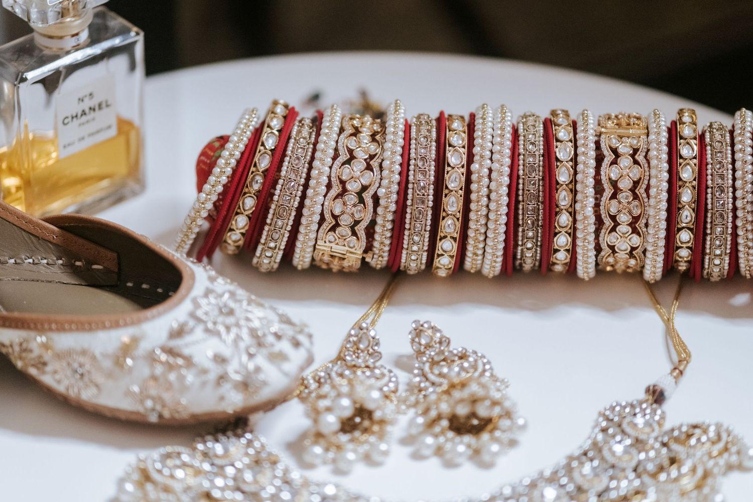 Red and gold Indian bridal bangles displayed with embellished shoes and jewelry beside Chanel perfume.