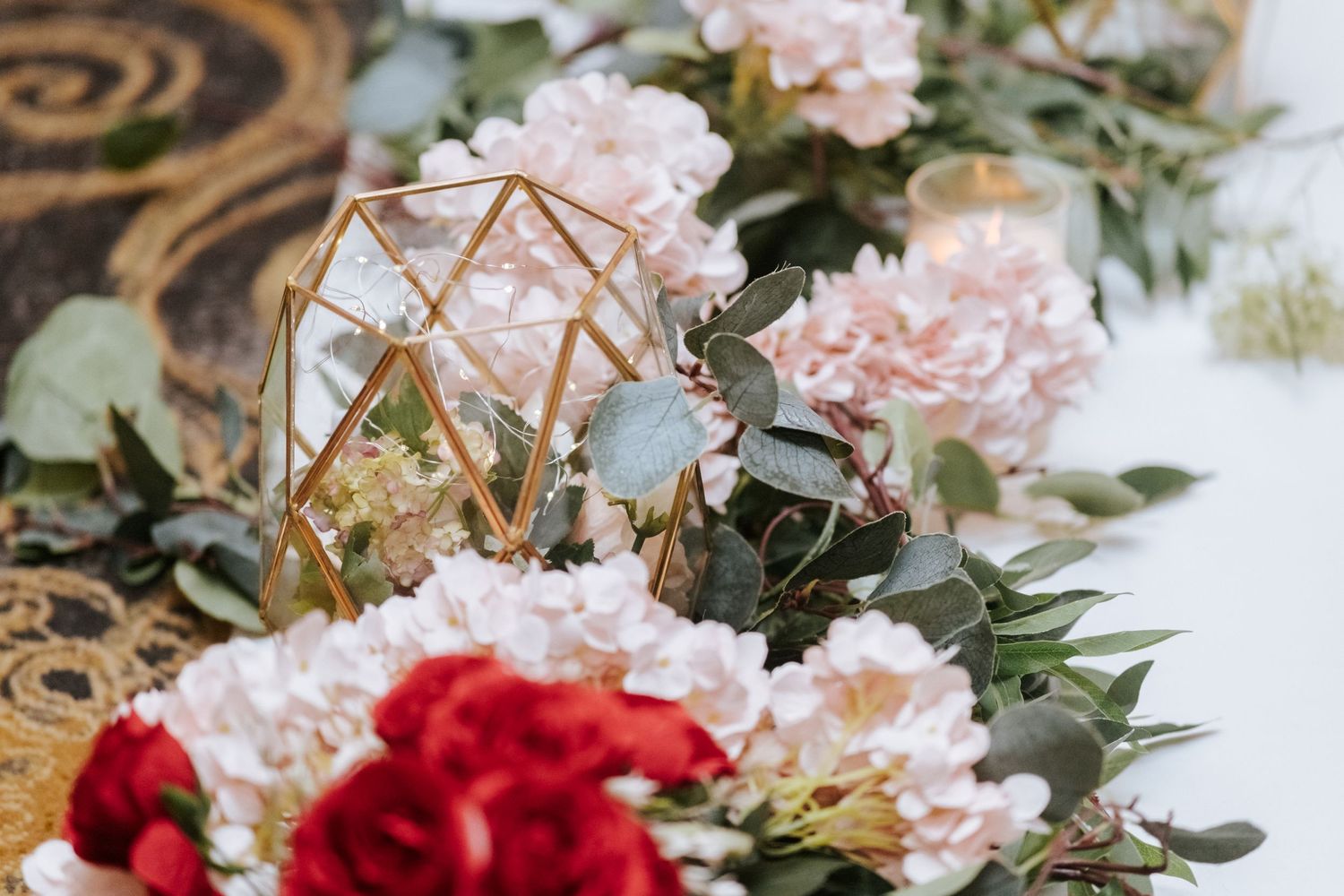 Gold geometric terrarium centerpiece surrounded by blush pink hydrangeas and red roses on a wedding table.