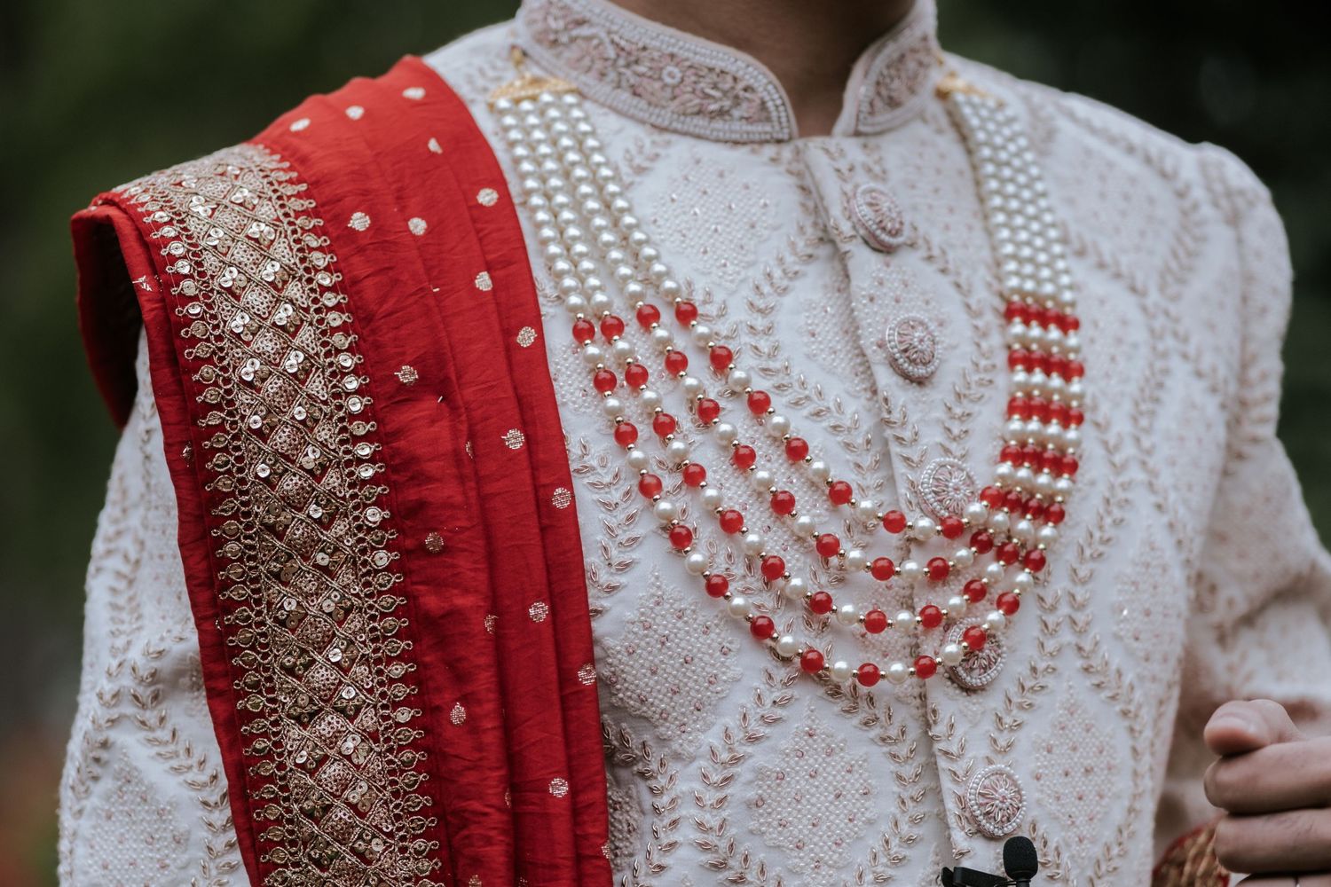 Close-up of Indian groom in white sherwani adorned with red dupatta and layered pearl and coral necklace.