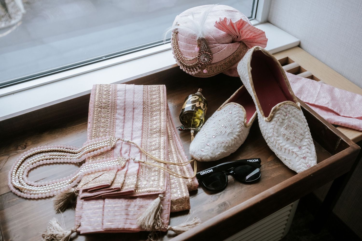 Indian groom wedding accessories including white juttis, pink turban, pearl necklace, sunglasses, and fabric on wooden tray.