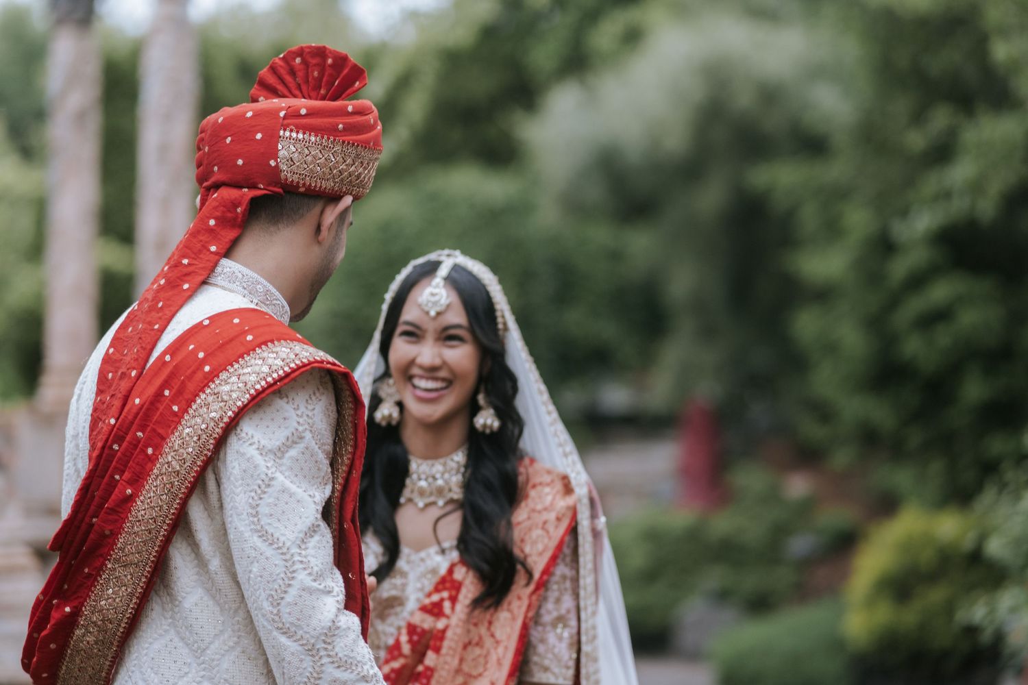 Indian bride and groom share a joyful first look moment outdoors in traditional red and white wedding attire.