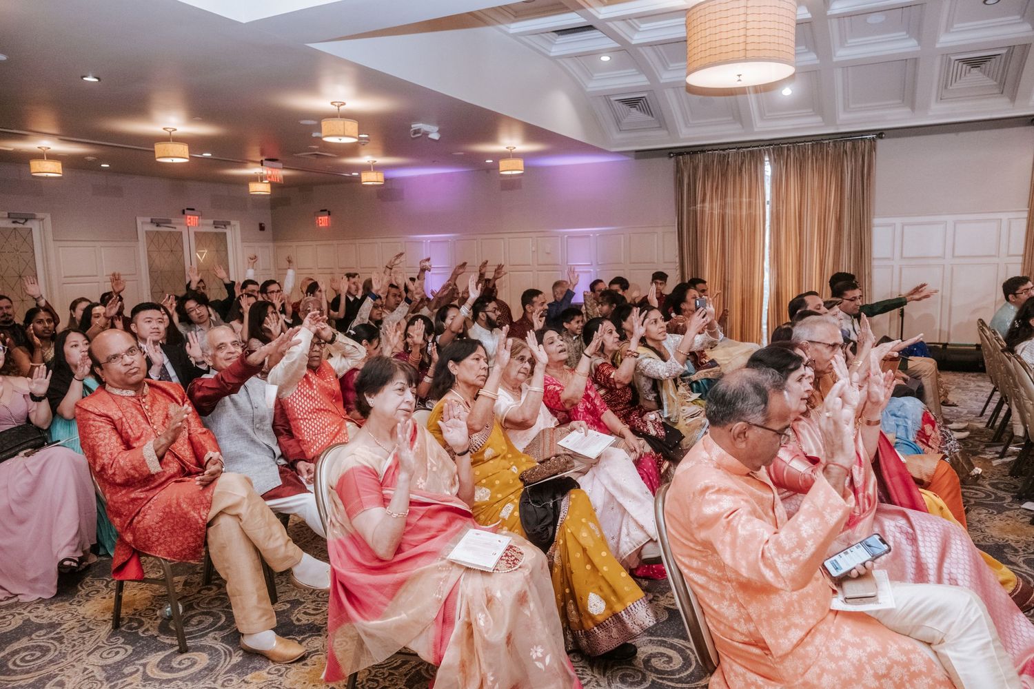 Large crowd of guests celebrating and cheering at a vibrant Indian wedding reception in a decorated ballroom.