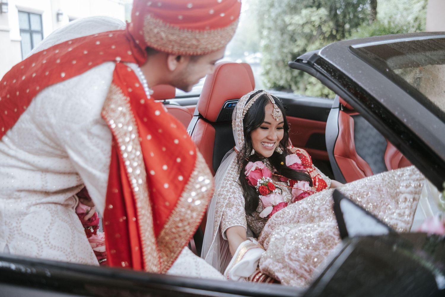 Indian groom in red sherwani leans into convertible to greet smiling bride in traditional wedding attire.