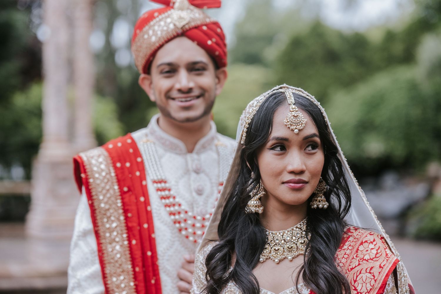 Indian bride and groom in red and white traditional wedding outfits posing outdoors.