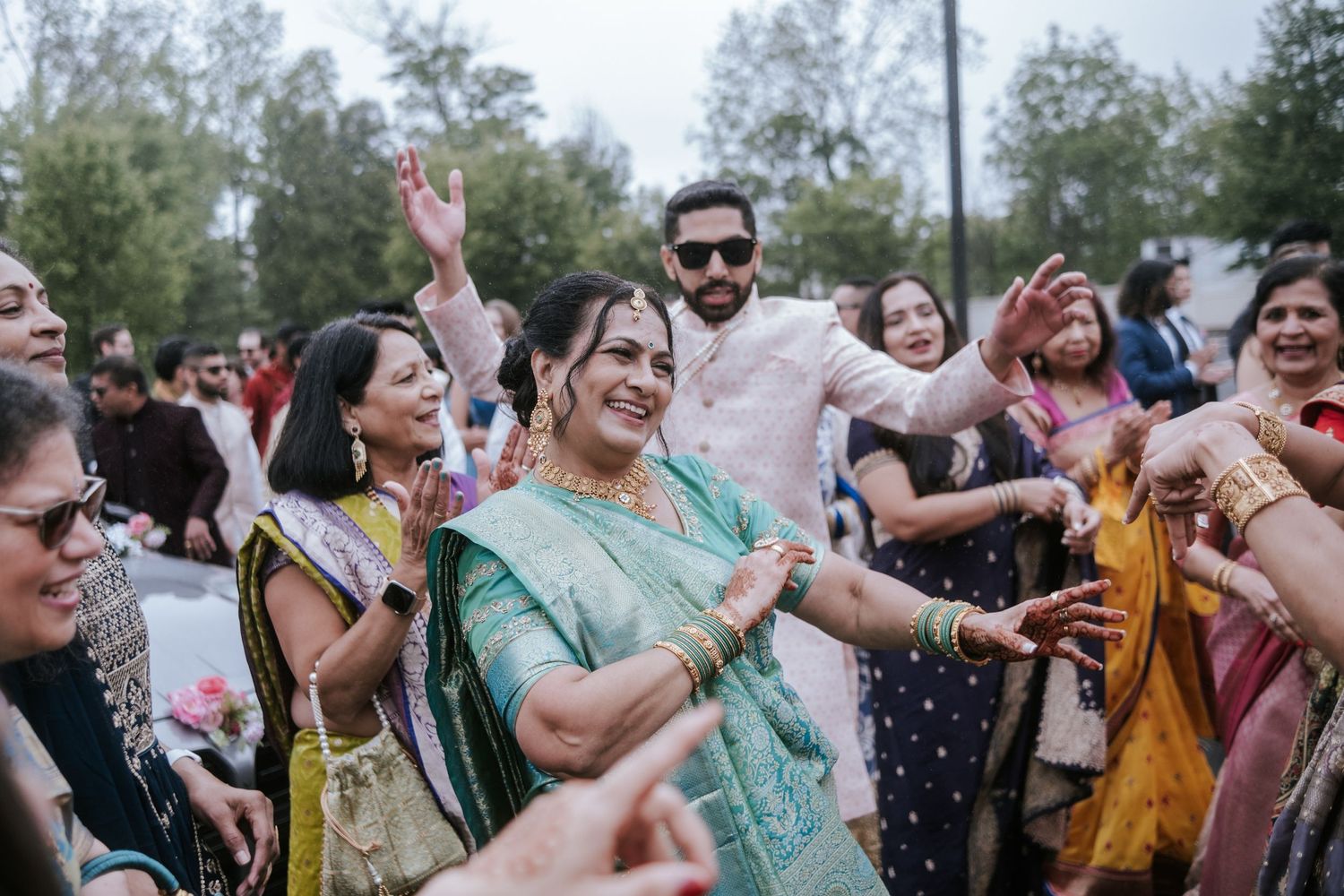 Woman in teal saree dances joyfully with guests at an Indian wedding celebration outdoors.