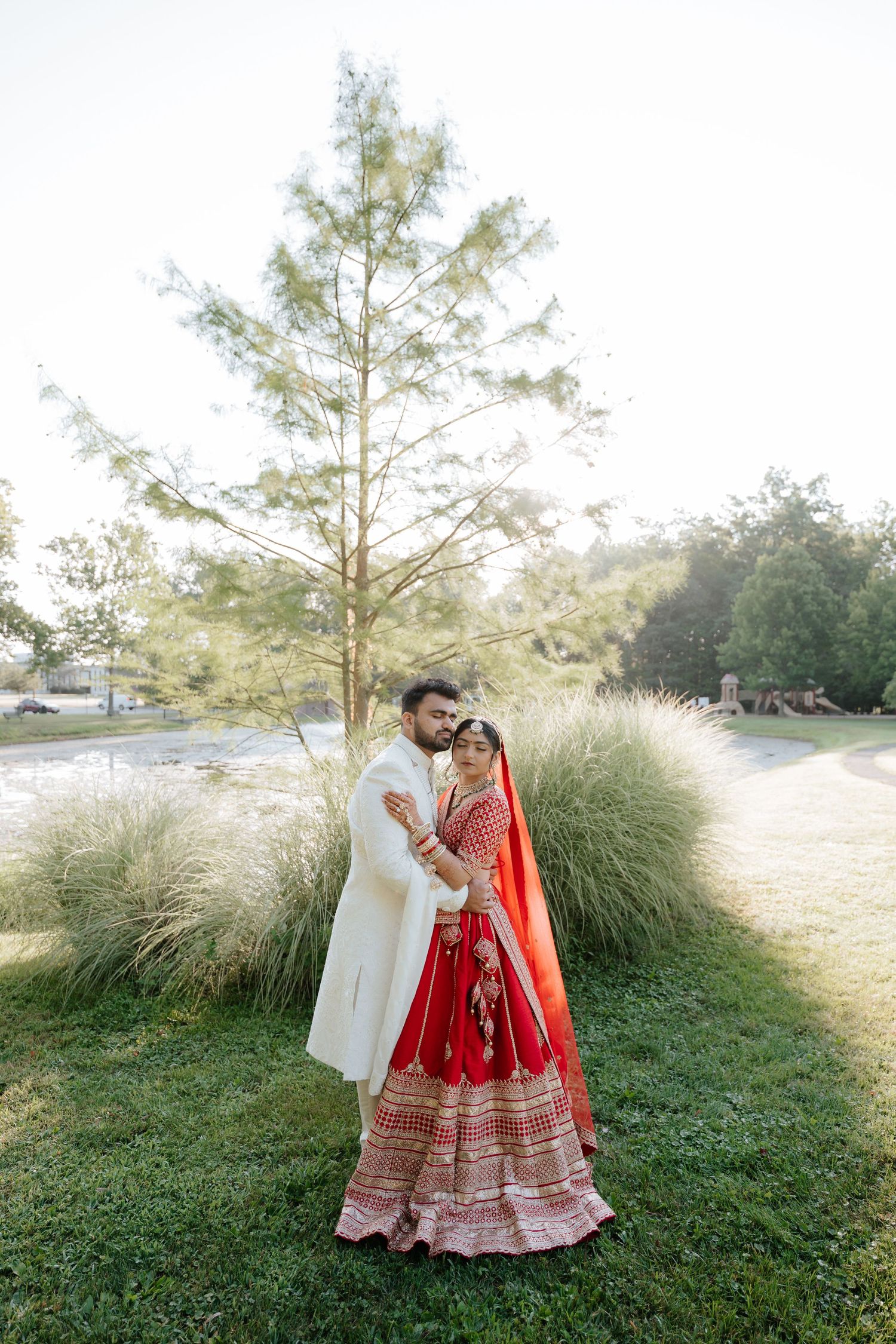 Indian bride and groom in red lehenga and white sherwani embracing near tall grasses by a pond.