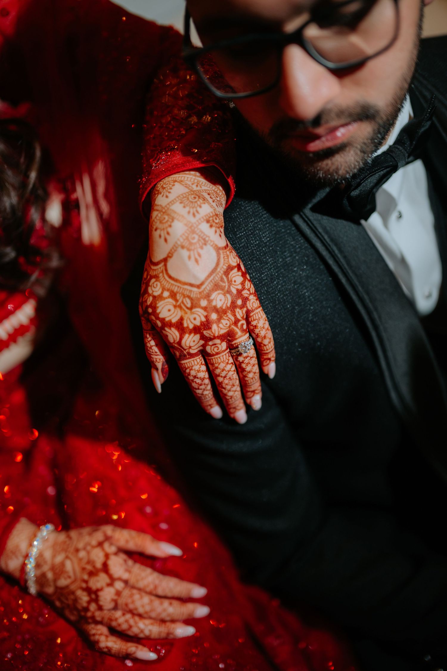 Close-up of intricate bridal mehndi on bride's hand resting on groom's black tuxedo jacket.