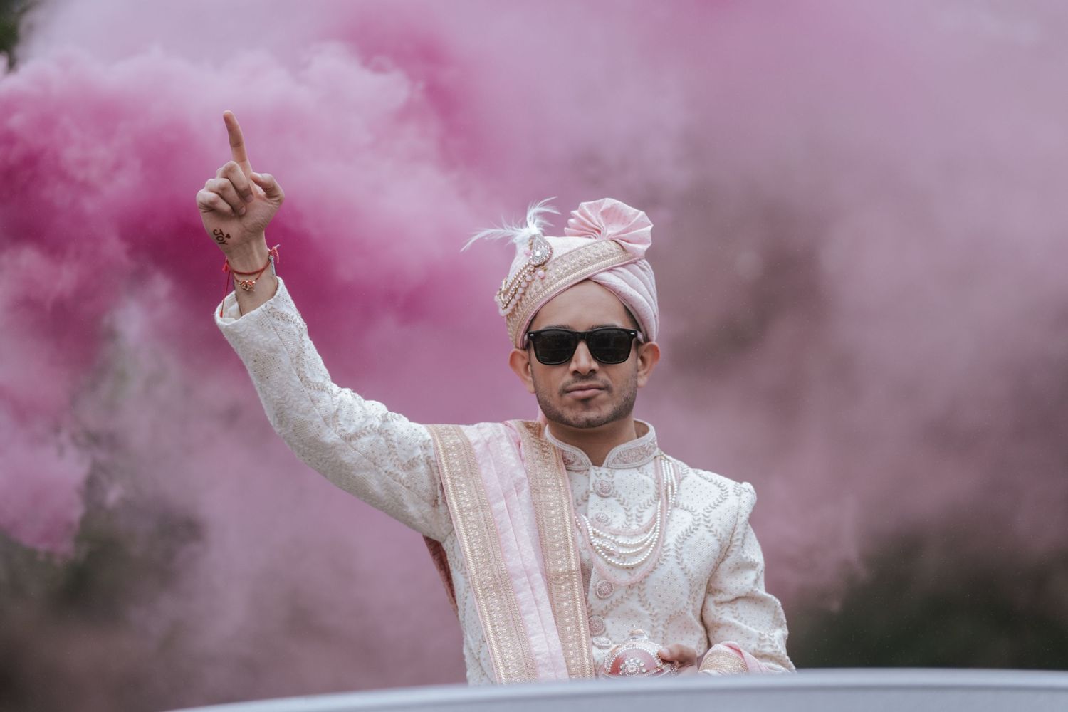 Groom in white sherwani and turban posing with pink smoke during Indian wedding baraat.