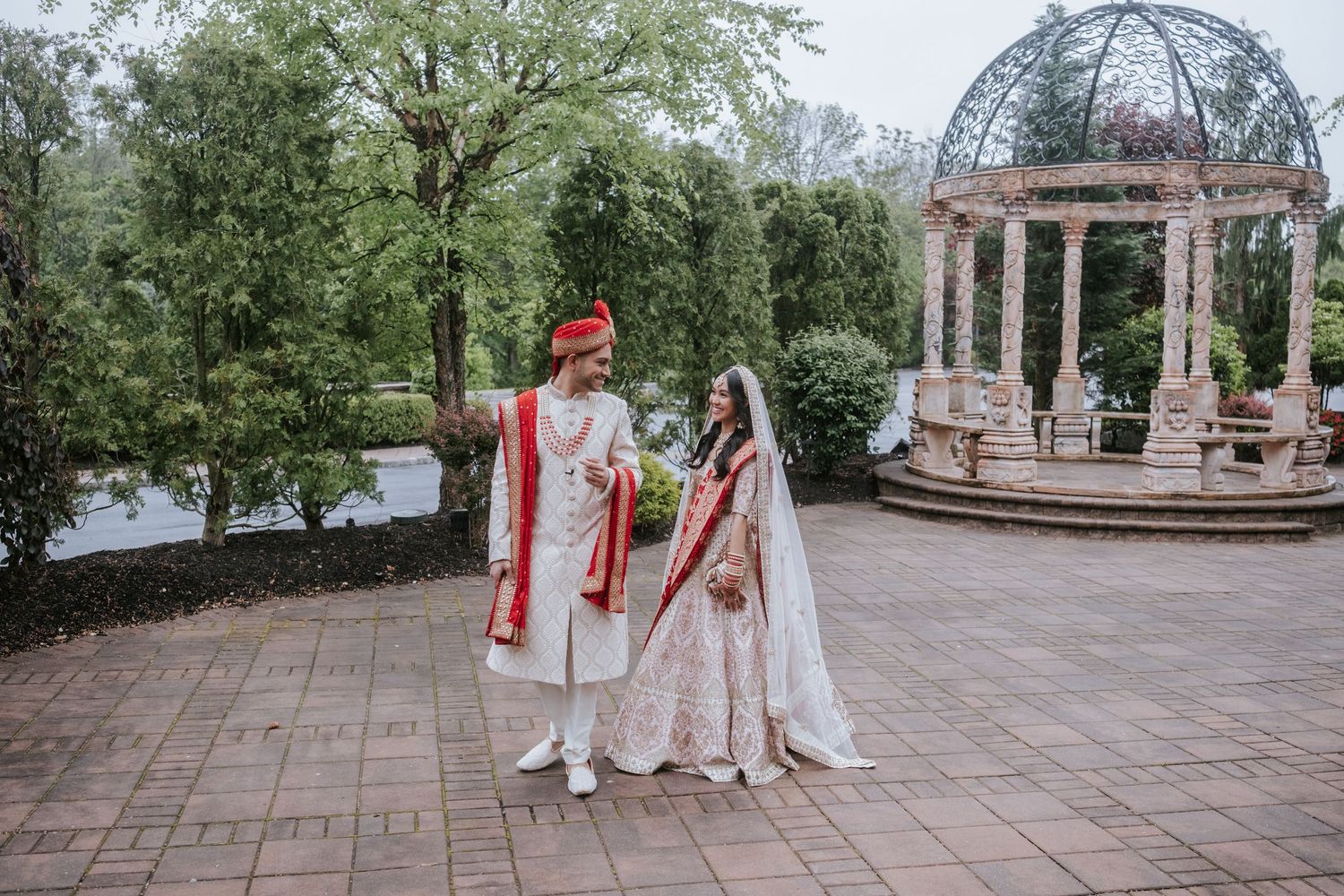 Indian couple in red and white wedding attire walking together near elegant stone gazebo.