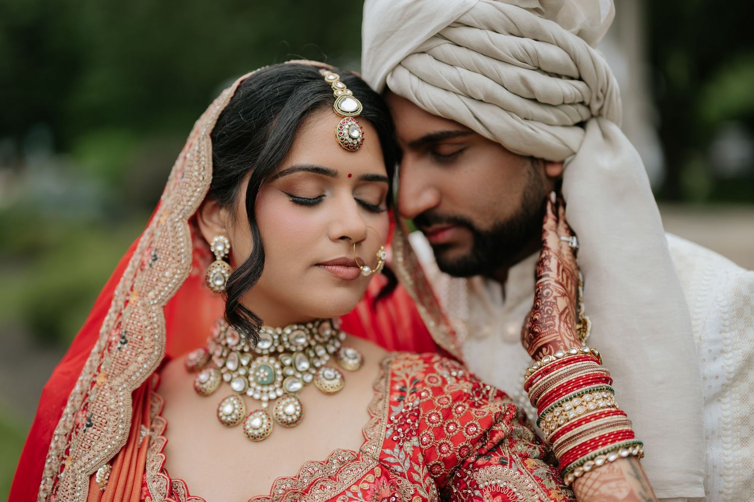 Indian bride in red lehenga with kundan jewelry and groom in white turban sharing tender moment.