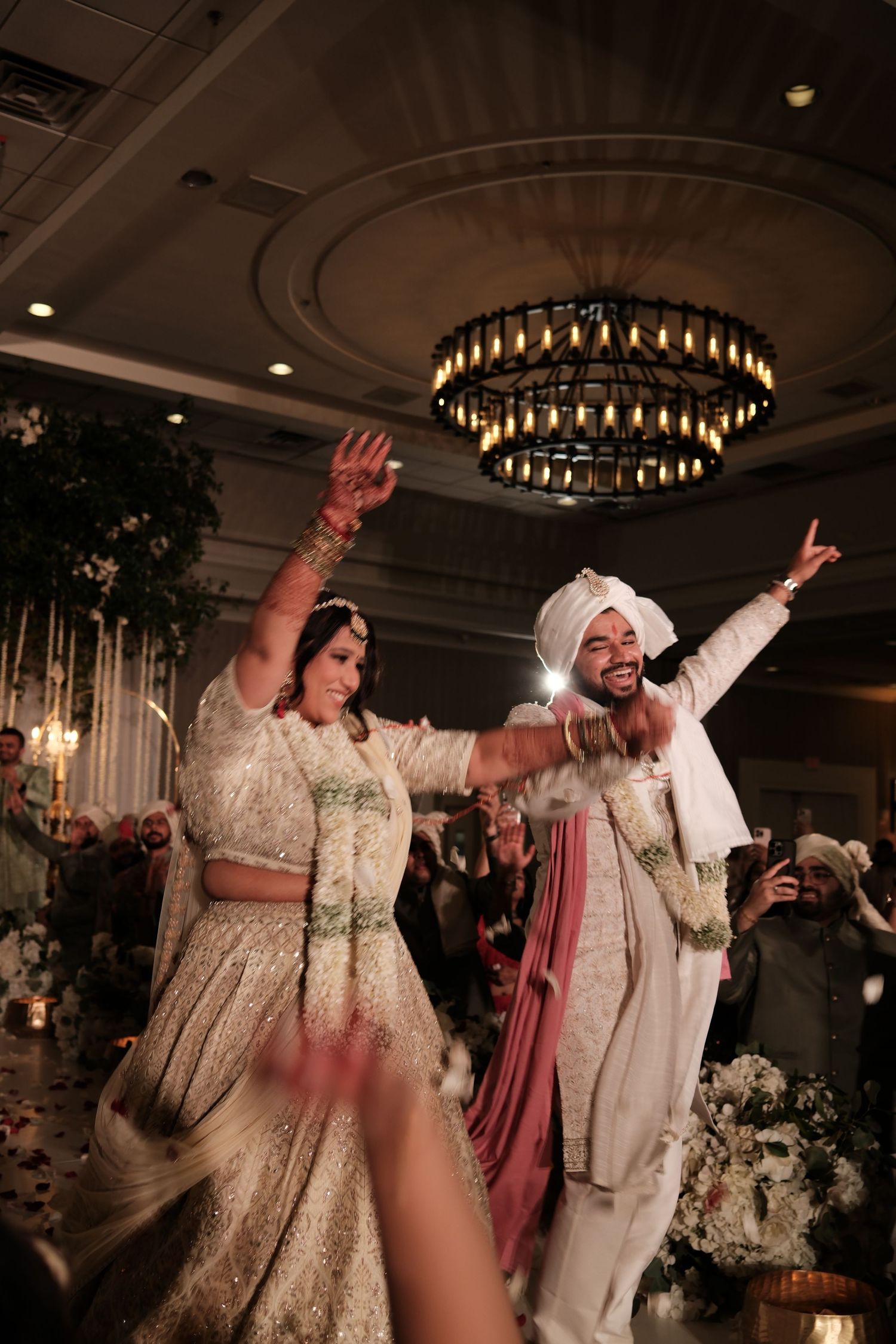 Indian newlyweds in ivory and white traditional attire dance joyfully at elegant wedding reception.