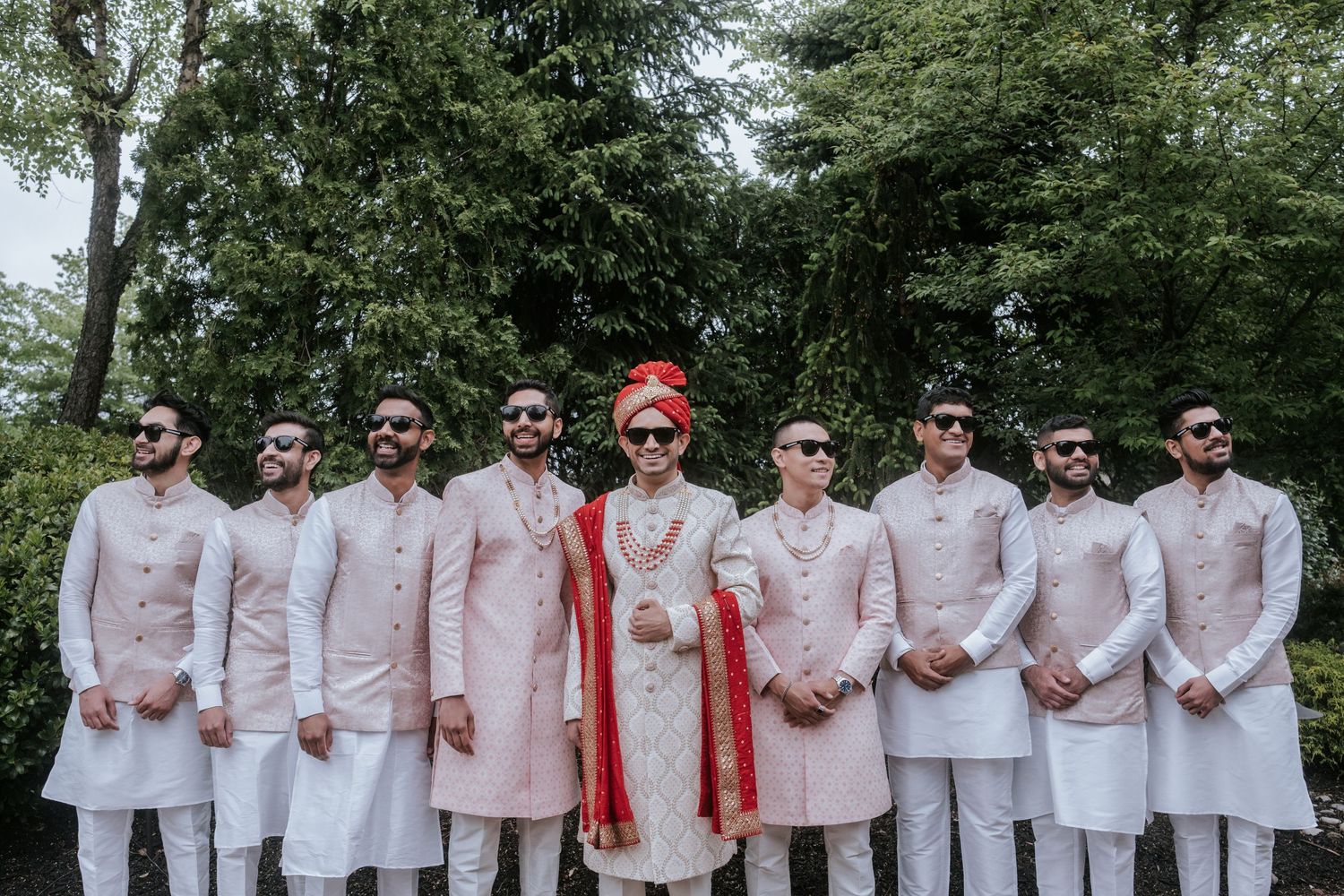 Indian groom in ornate red and white sherwani stands with groomsmen in matching pink kurtas outdoors.