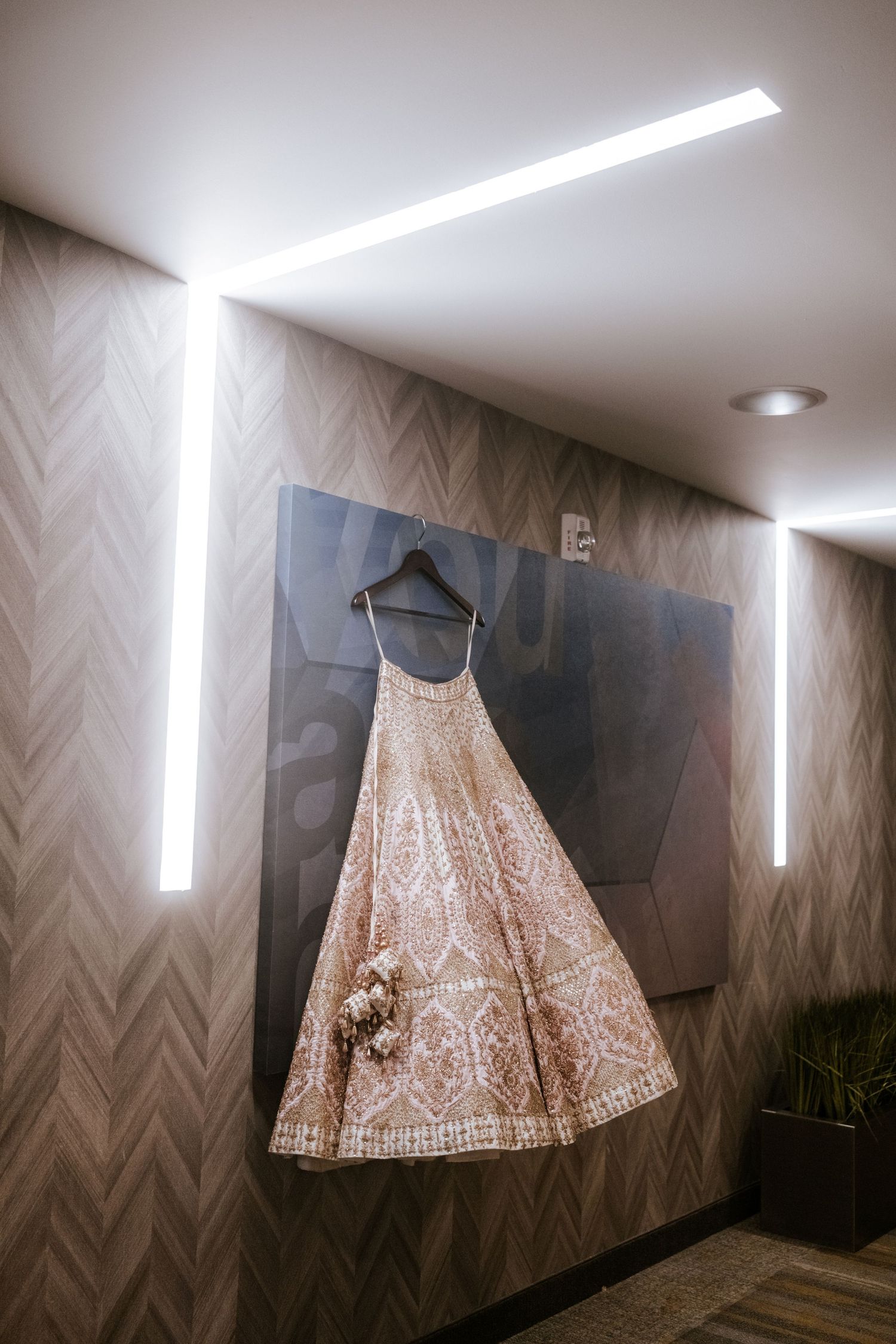Elegant ivory and gold embroidered bridal lehenga hanging on a hanger against a modern wood-paneled wall.