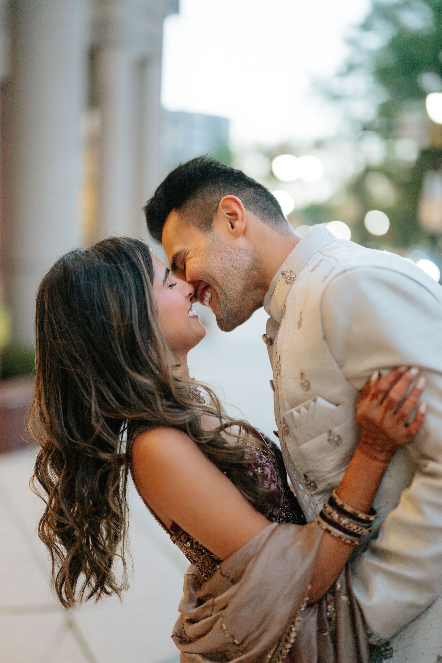 Indian couple in traditional attire sharing a romantic moment, touching foreheads and smiling outdoors.