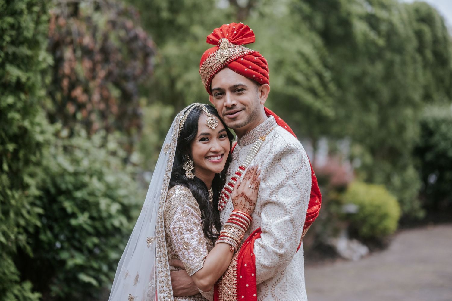 Indian bride and groom posing together outdoors, bride in white lehenga with veil, groom in white sherwani and red turban.