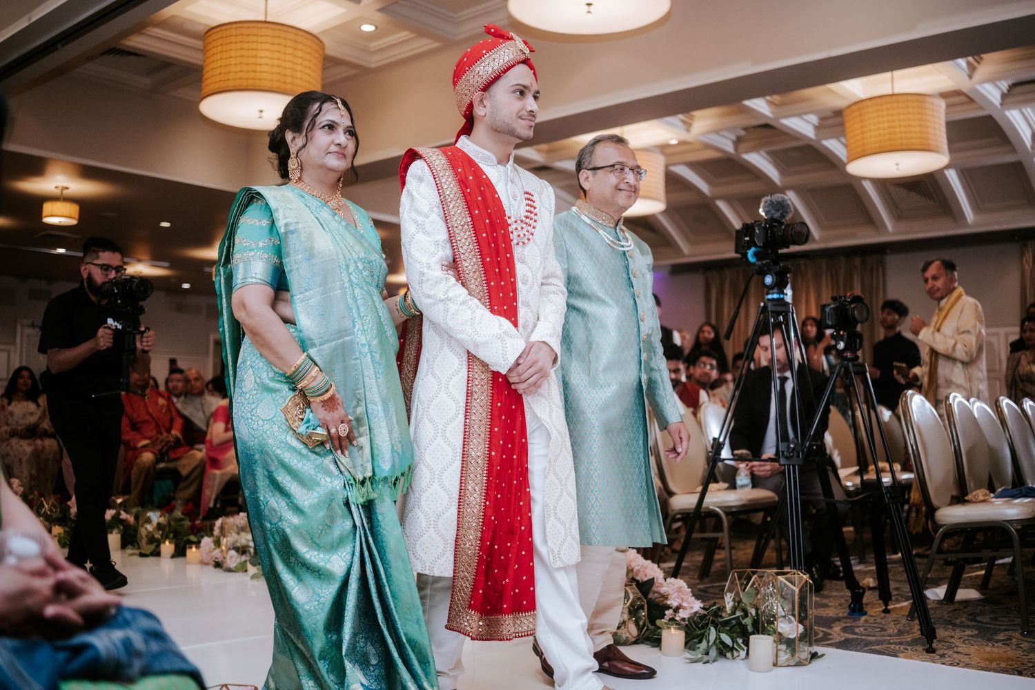 Indian groom in red and gold sherwani walks down aisle escorted by family at wedding ceremony.