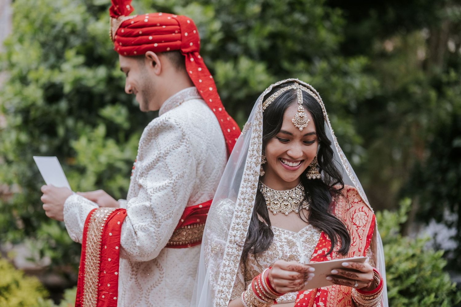 Indian bride smiling while reading vows, groom standing back-to-back with her in traditional red and white wedding attire.
