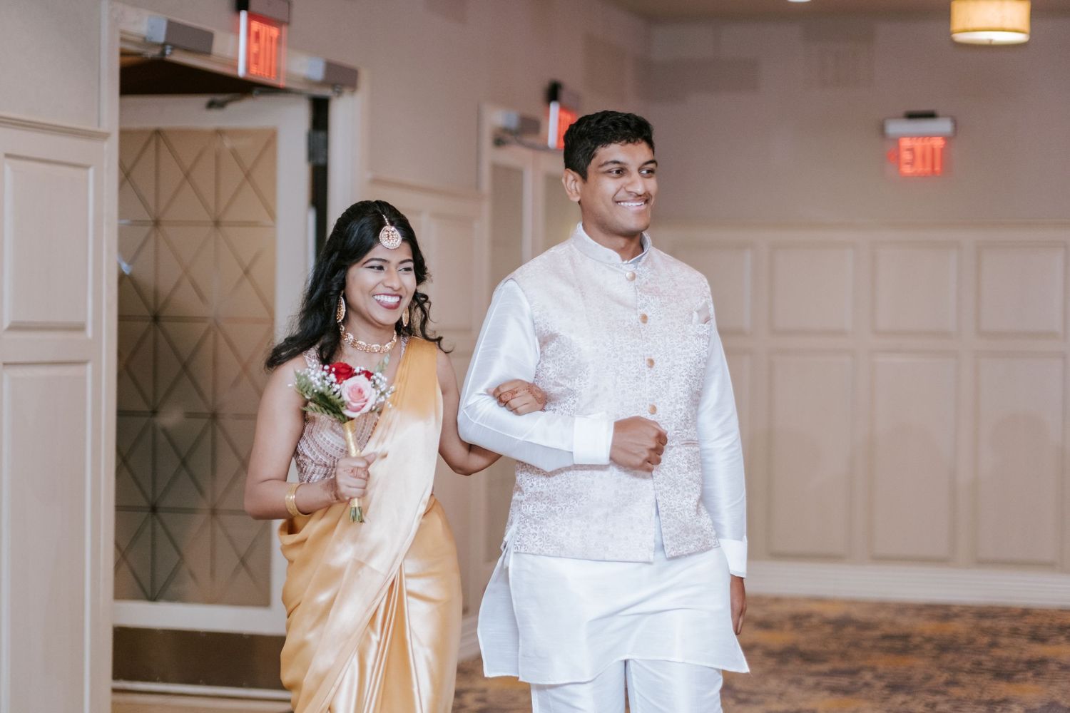 Bride in gold saree and groom in white sherwani walking into Indian wedding reception.