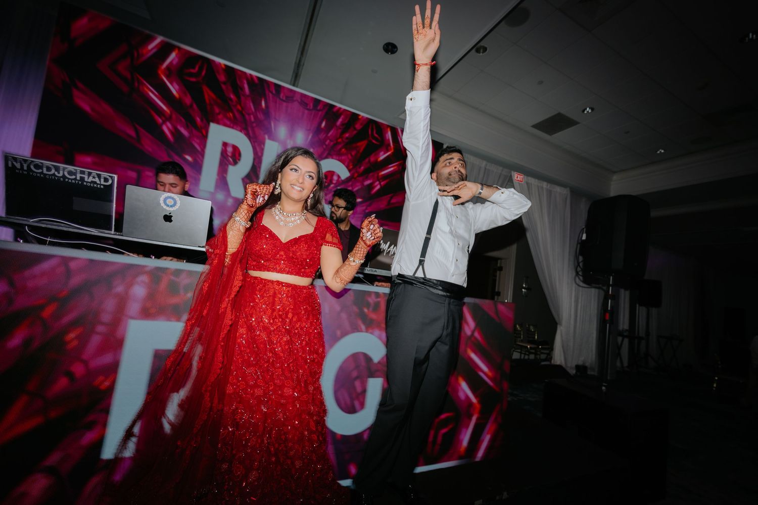 Indian couple in red and grey outfits dancing energetically on stage at a vibrant wedding reception.