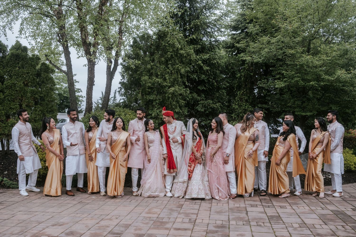 Indian wedding party in coordinated pink and gold traditional attire poses in large group outdoors.