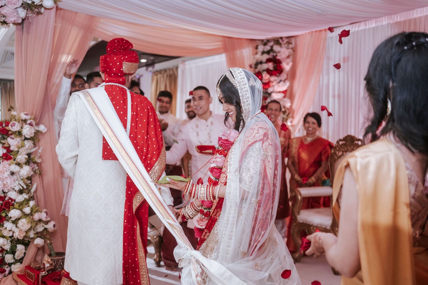 Indian bride and groom exchanging vows at a traditional Hindu wedding ceremony with red and white floral mandap.