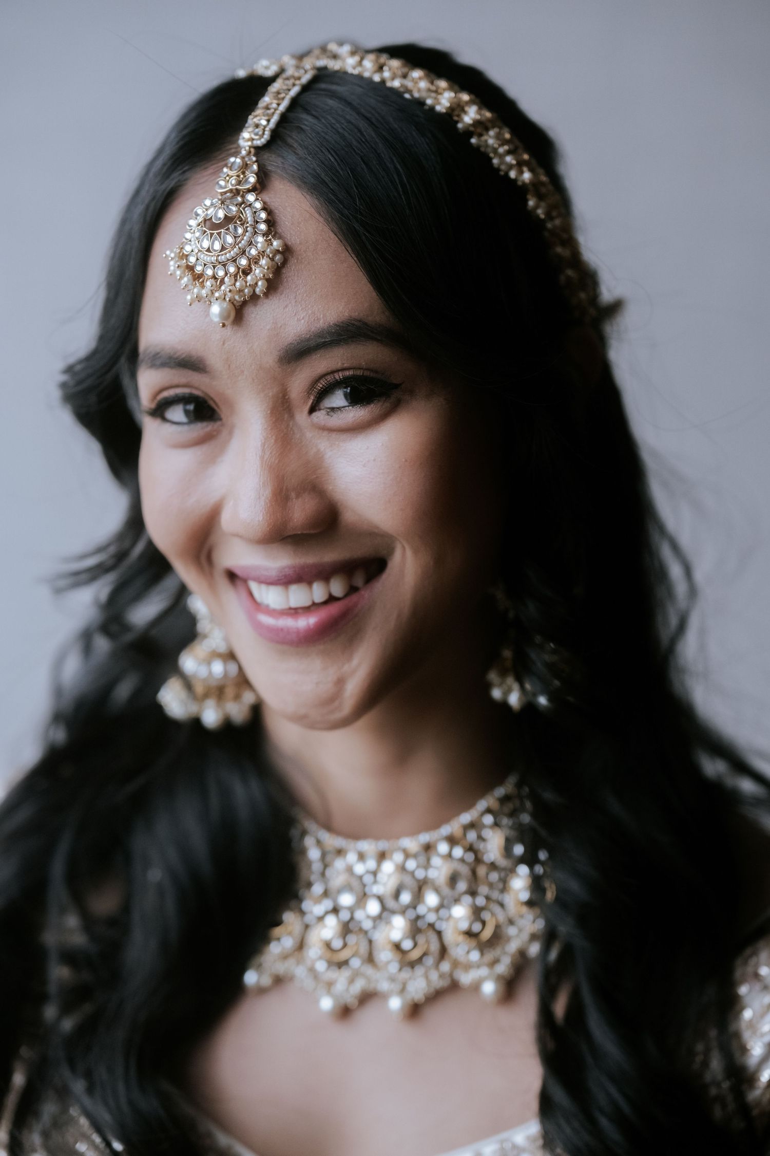 Smiling Indian bride wearing ornate gold maang tikka, chandelier earrings, and embellished necklace.
