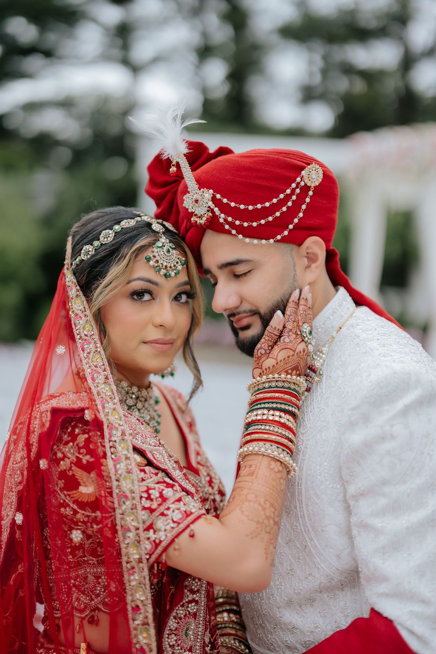 Indian bride in red lehenga with mehndi hands cradles grooms face in intimate wedding portrait.