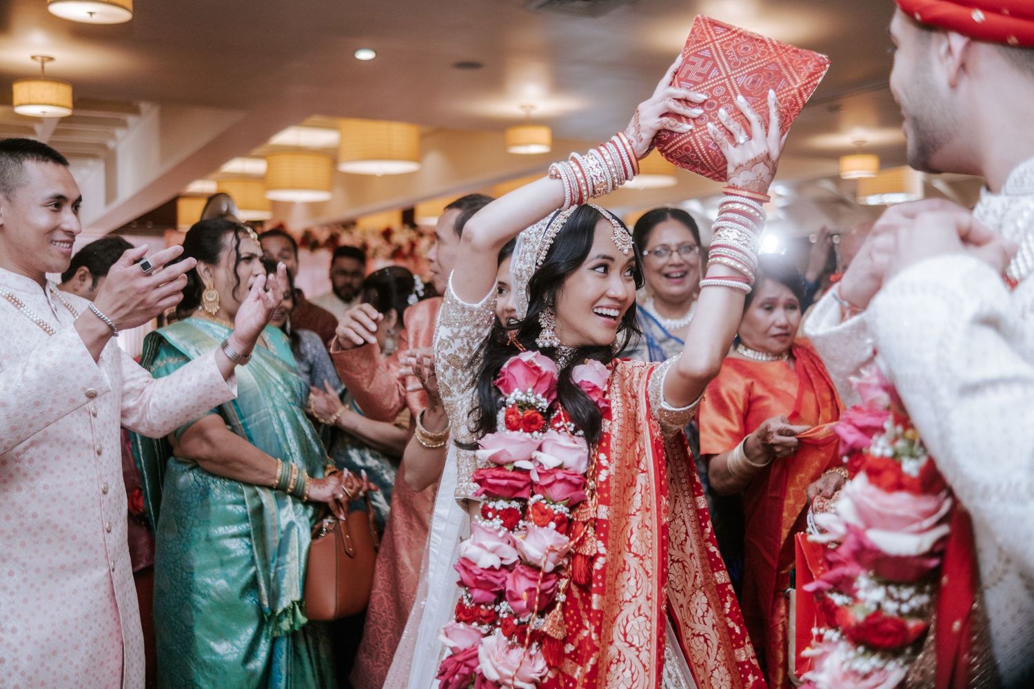 Smiling Indian bride holding red book overhead walks through cheering guests during wedding procession.