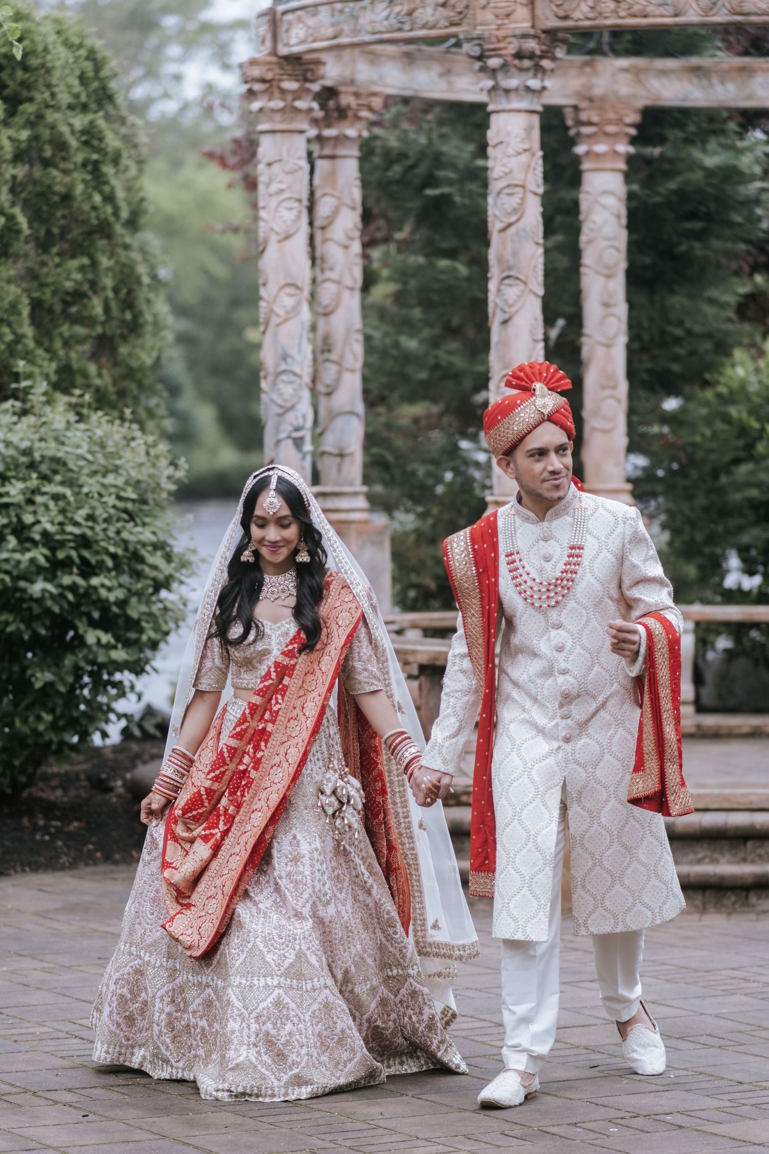 Indian bride and groom walking hand in hand in traditional wedding attire near ornate stone gazebo outdoors.