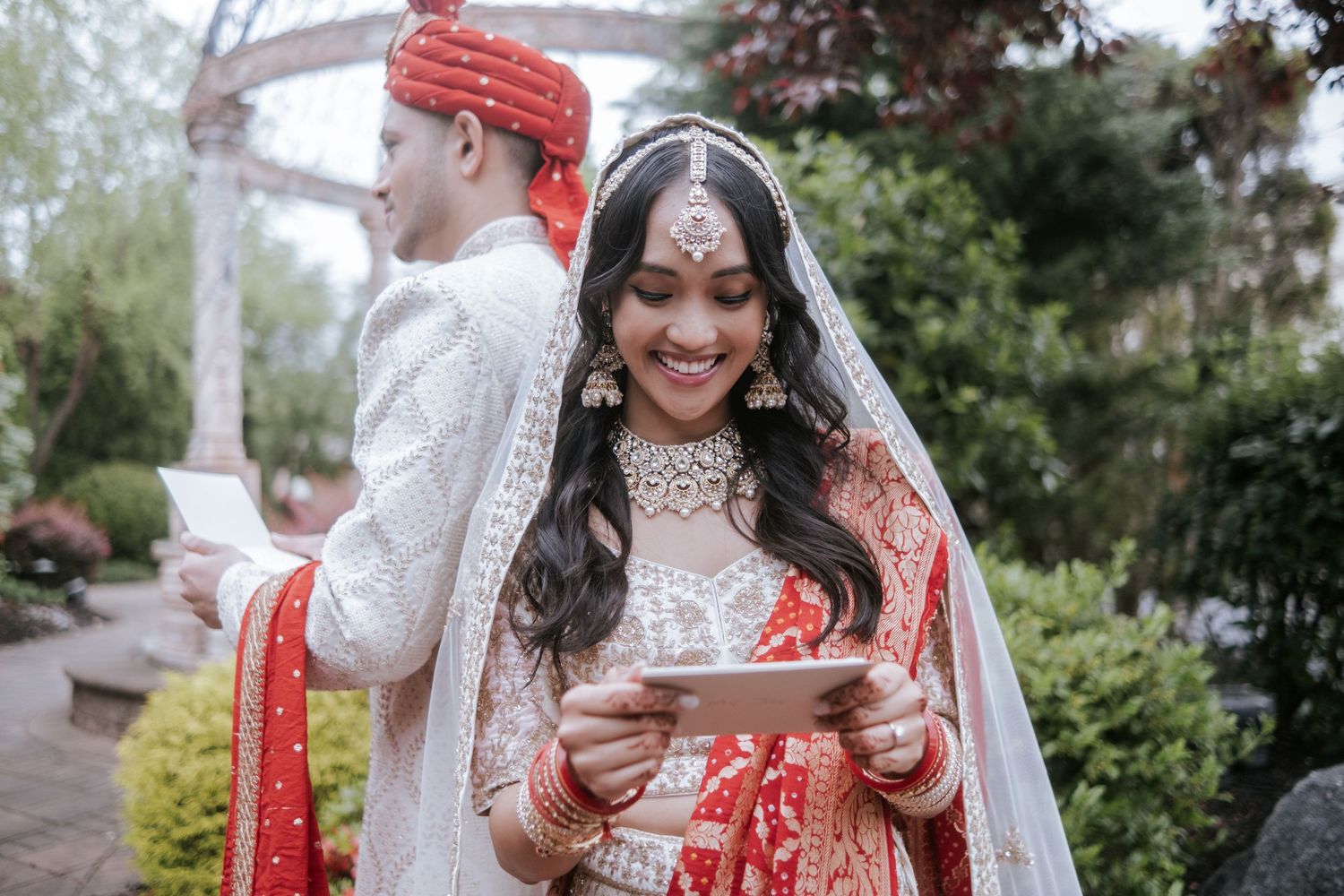 Beautiful Indian bride in red and gold lehenga smiles at phone while groom stands behind in garden setting.