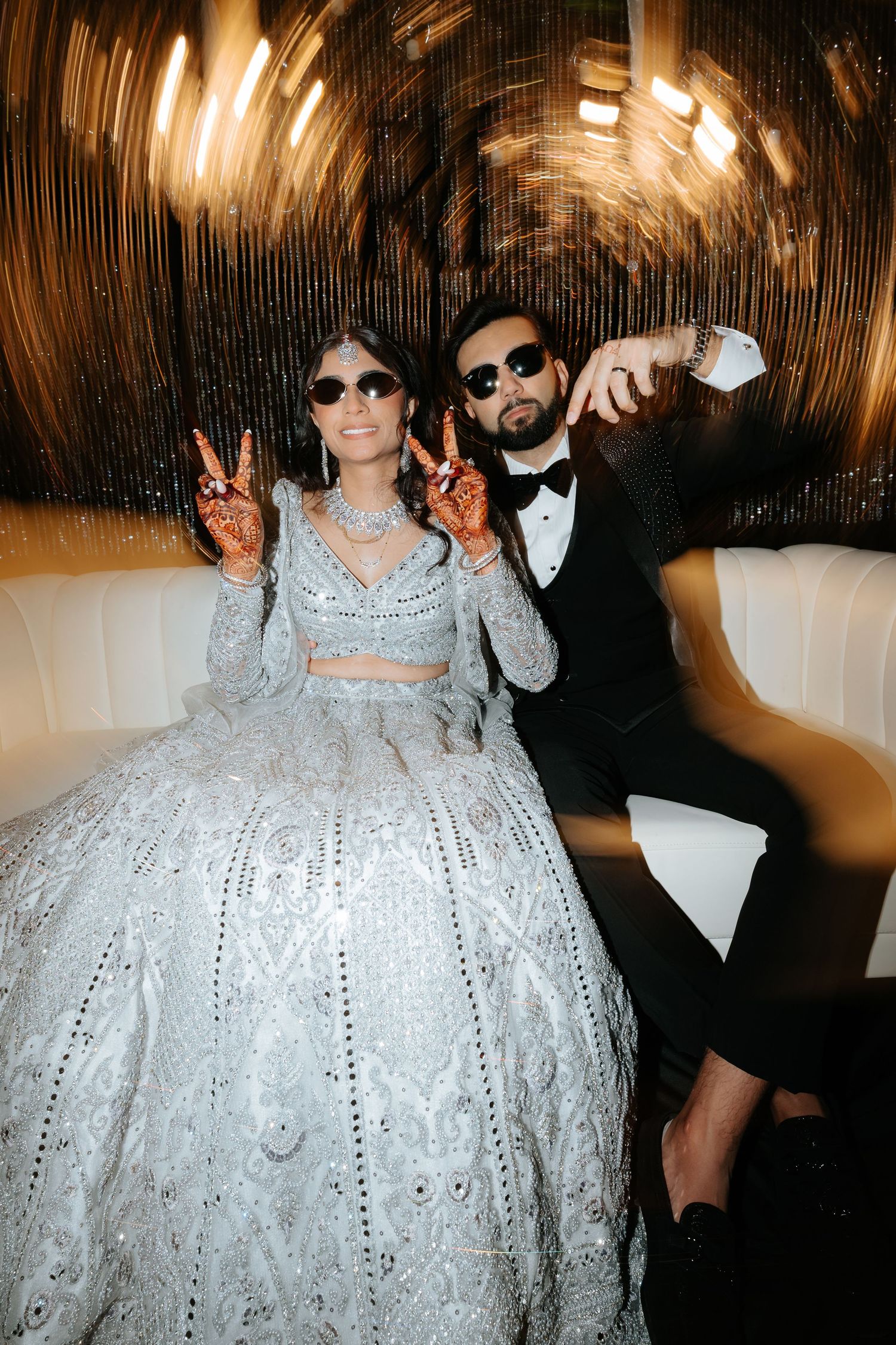 Indian bride in silver lehenga and groom in black tuxedo posing coolly with sunglasses at reception.