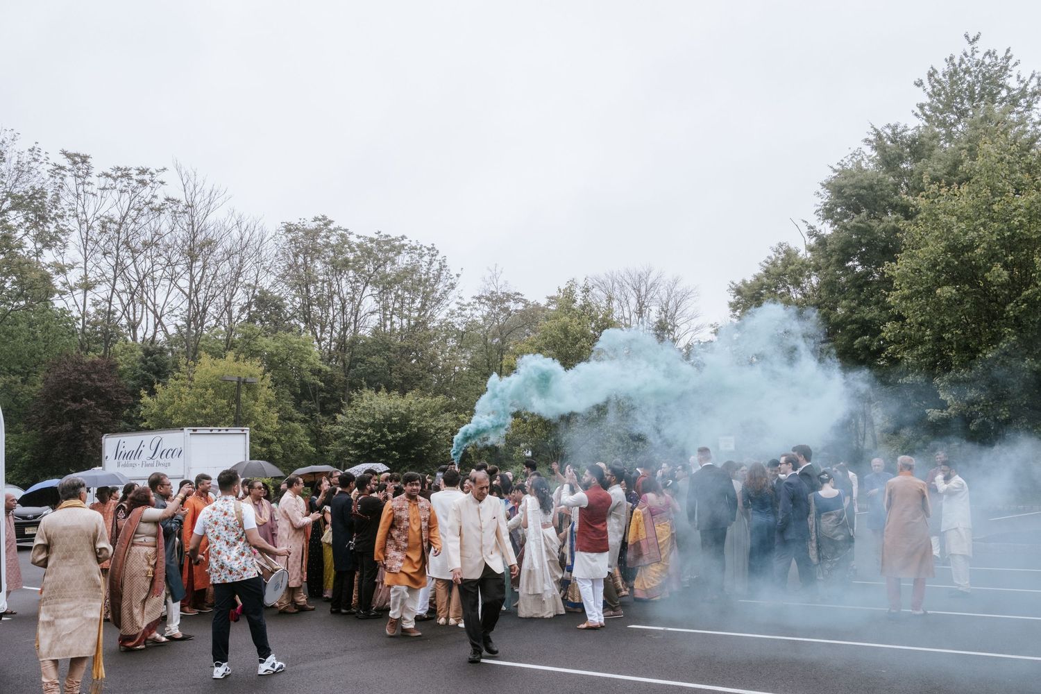 Indian wedding baraat procession with colorful teal smoke bomb in an outdoor parking lot.