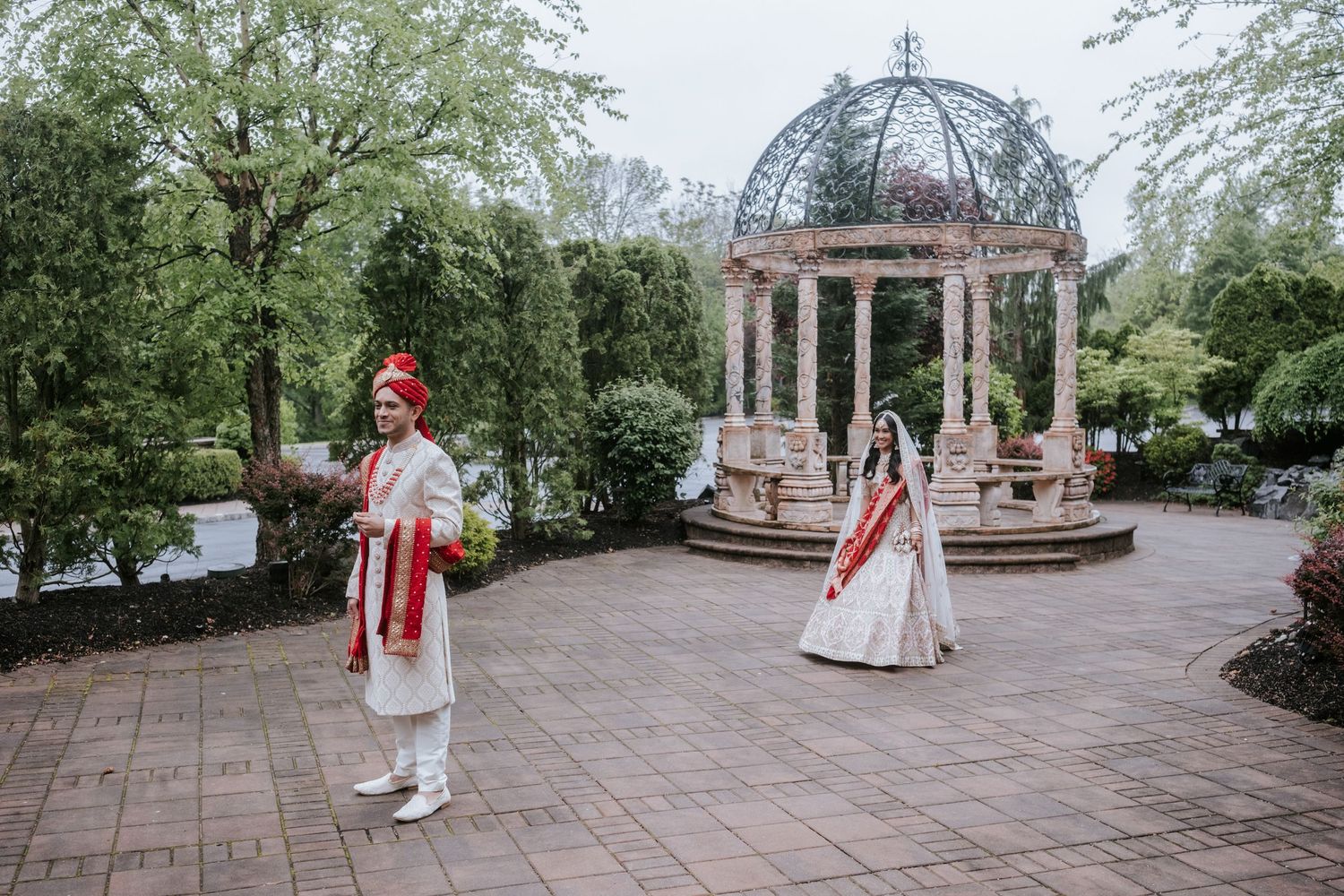 Indian bride and groom in traditional attire near stone gazebo in lush garden setting.