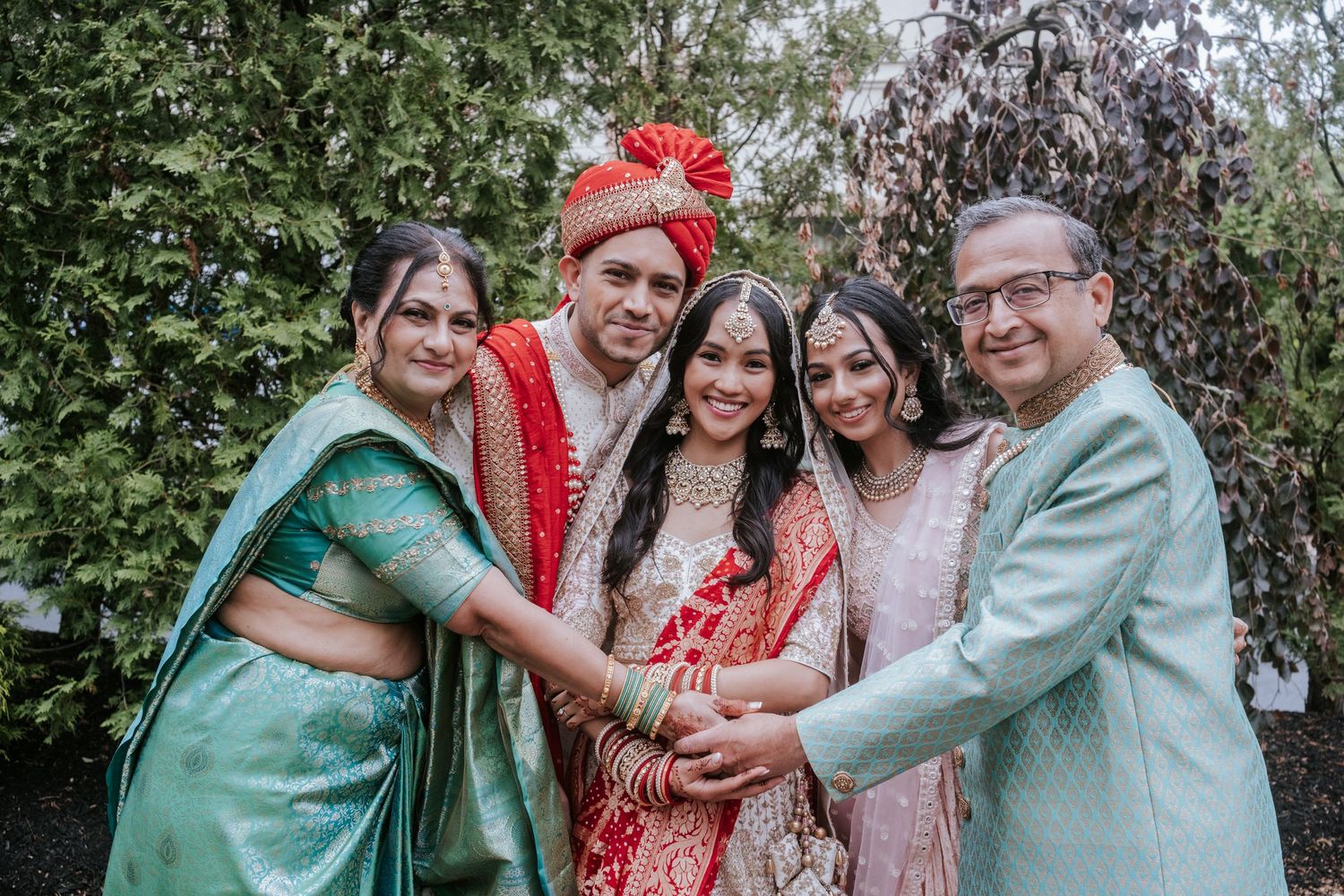 Indian wedding family portrait with bride, groom, and three relatives in traditional South Asian attire outdoors.