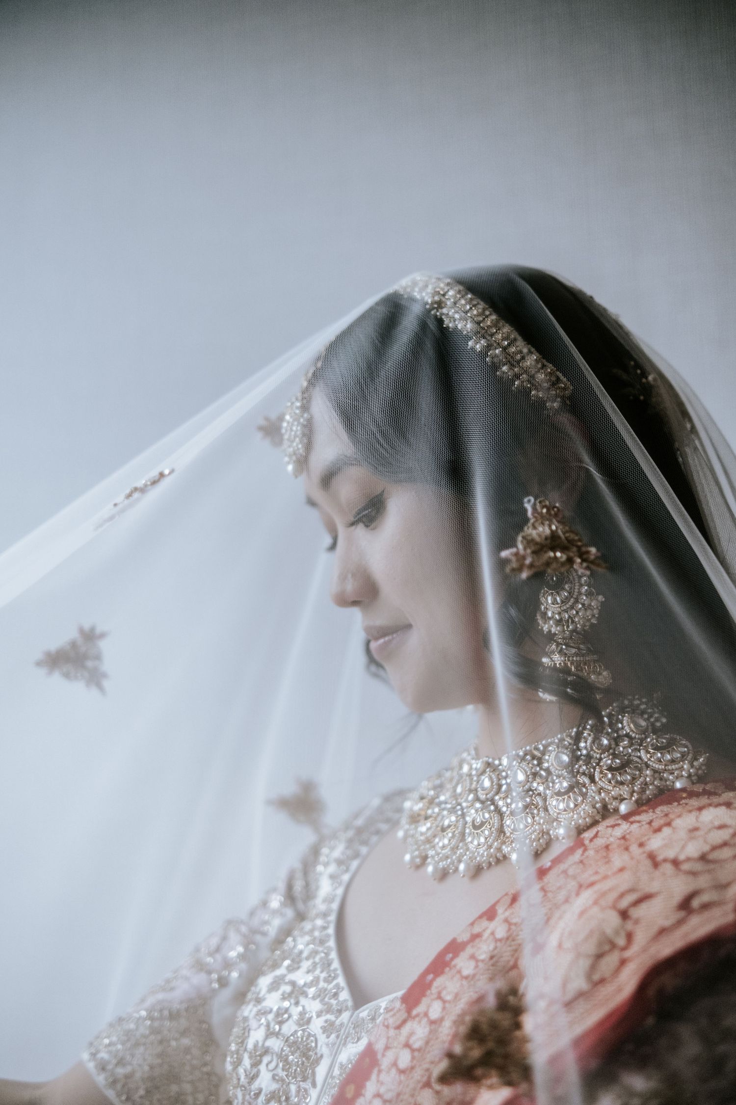 Indian bride draped in sheer veil looks down serenely in embroidered bridal lehenga.