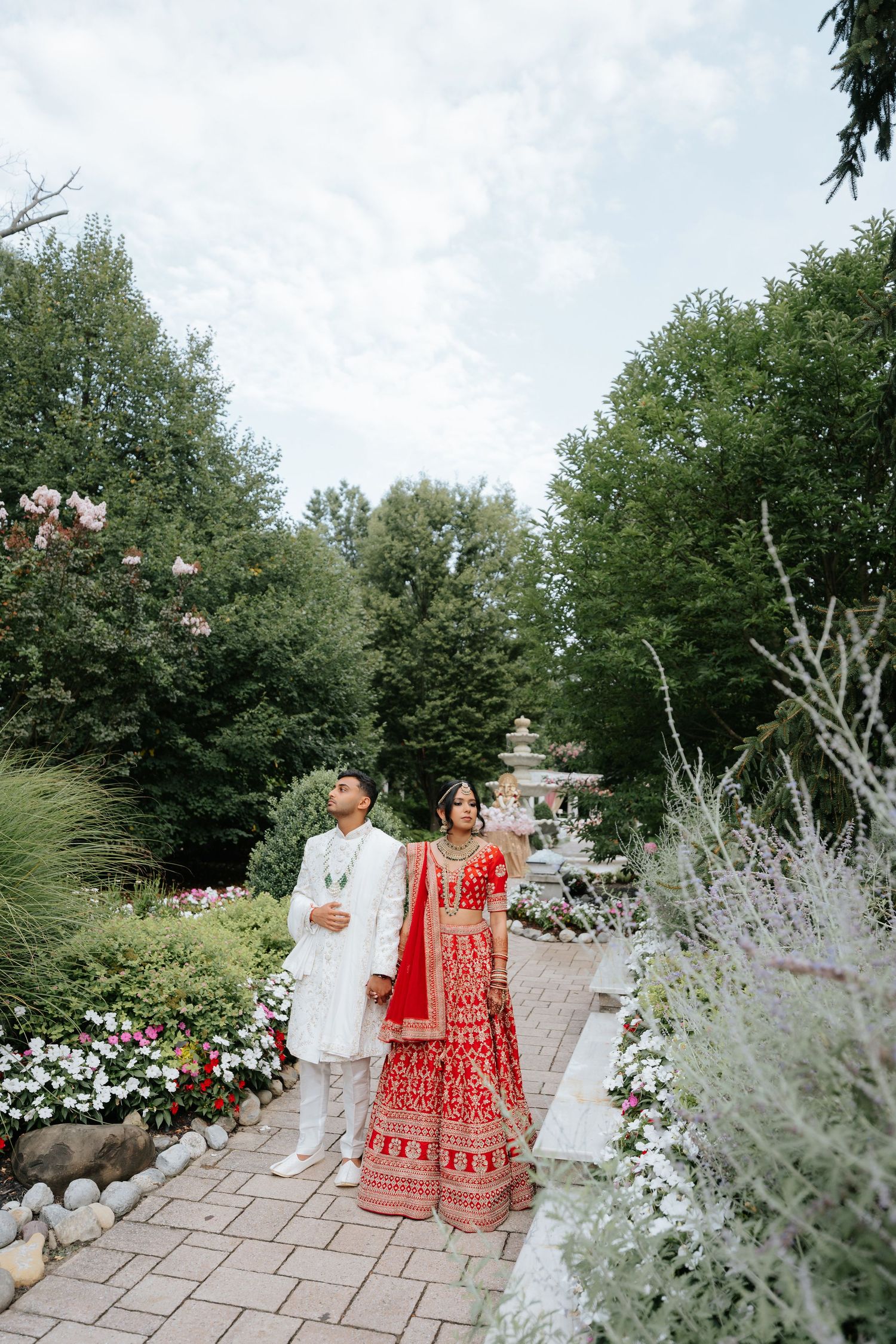 Indian bride and groom in traditional attire walking through a lush garden wedding venue.