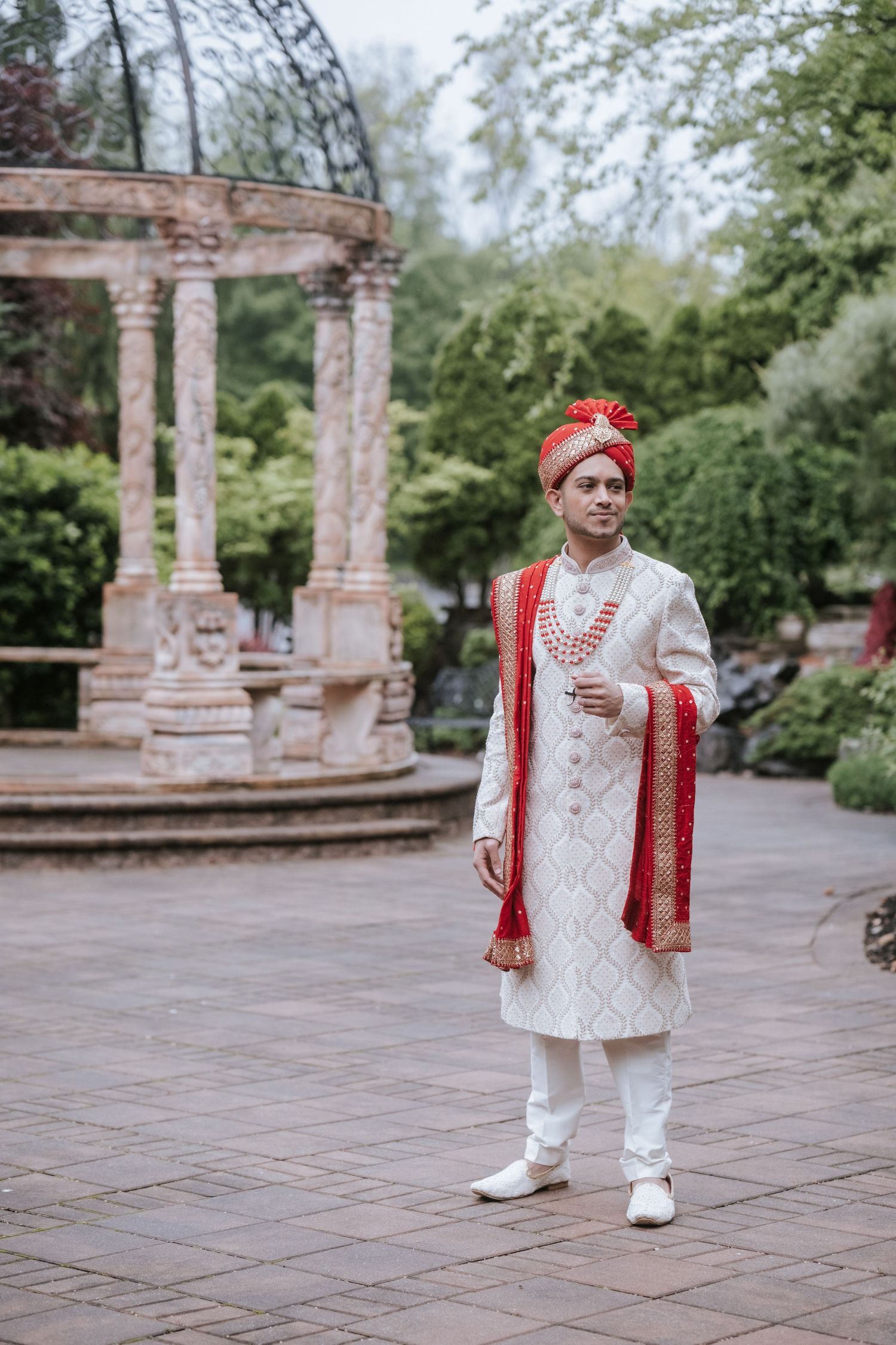 Indian groom in white and red sherwani with red turban stands confidently in lush garden near stone columns.