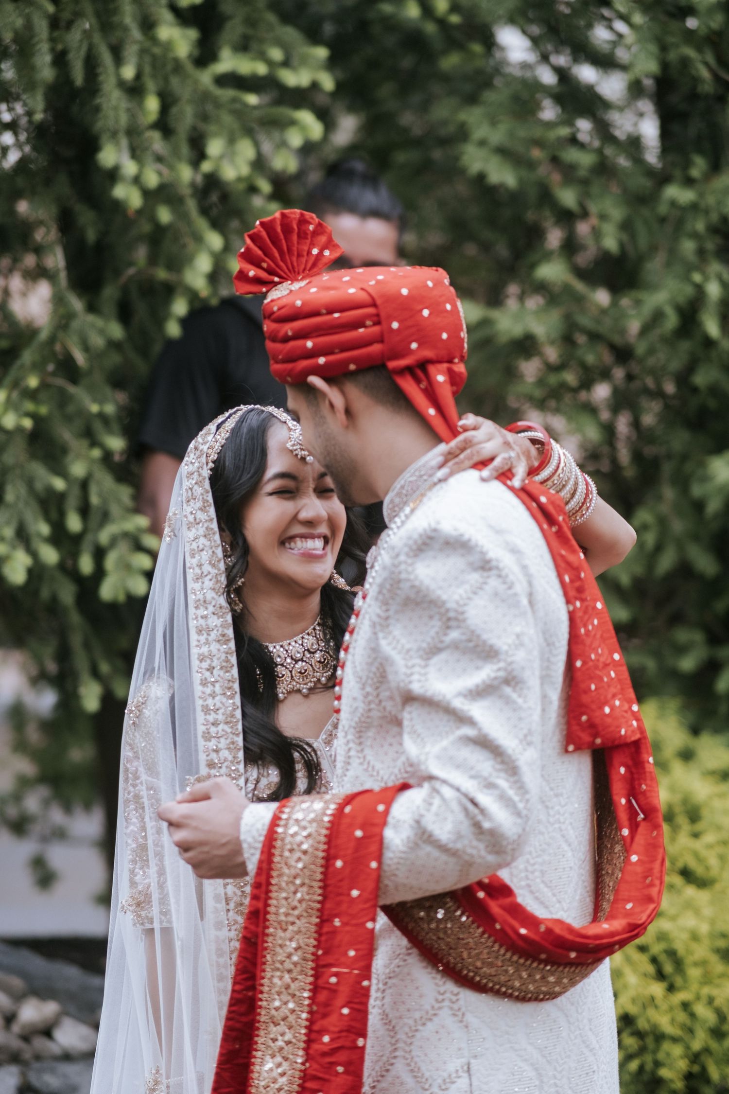 Joyful Indian bride laughing as groom in red turban whispers to her during outdoor wedding portrait session.