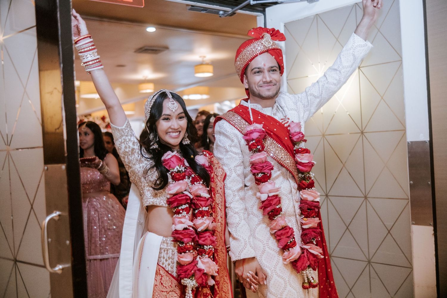 Happy Indian newlyweds raise arms in celebration wearing traditional red and white wedding attire with garlands.