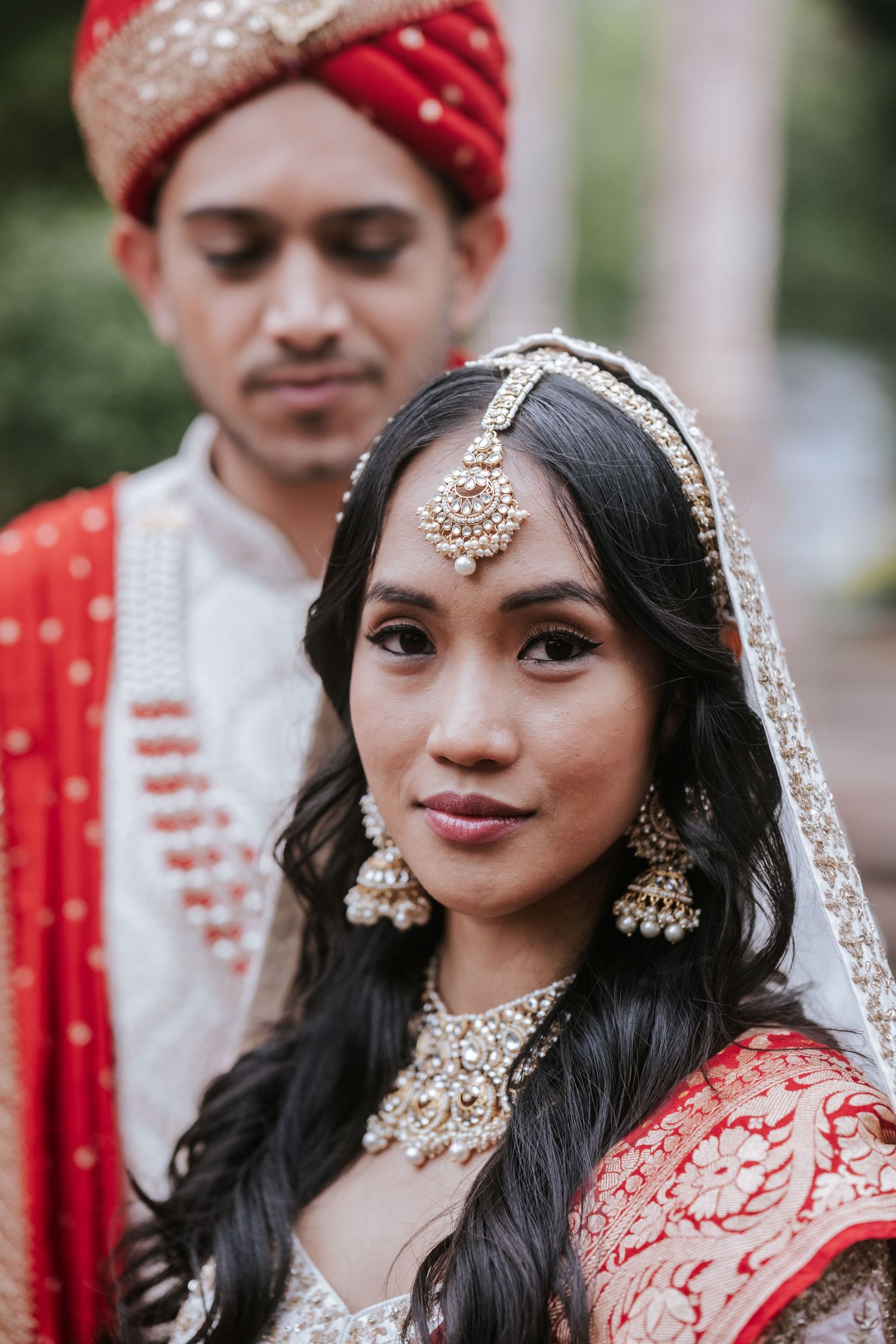 Indian bride in red and gold bridal jewelry with maang tikka and jhumkas, groom in red turban behind her.