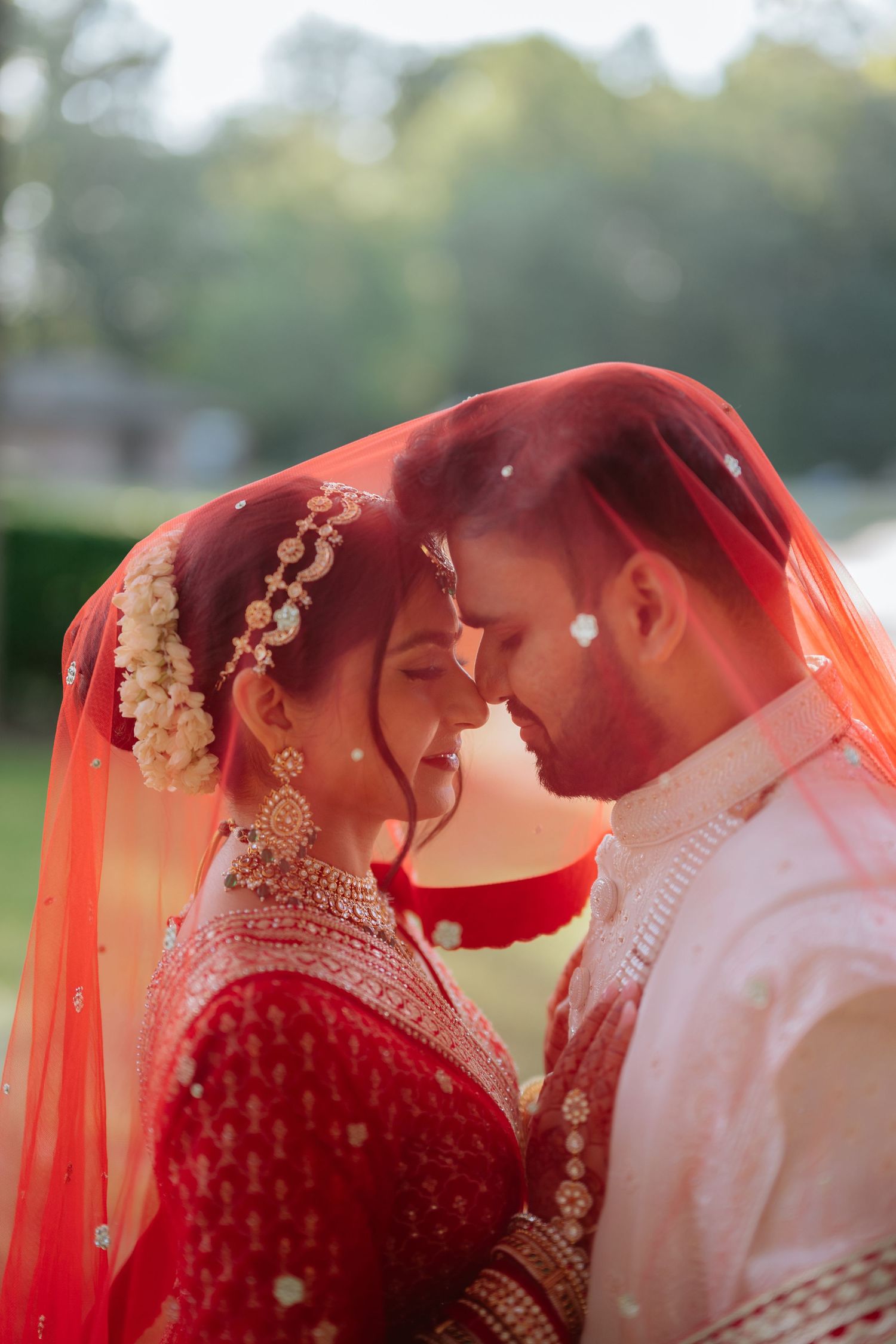 Indian bride and groom sharing intimate moment under red dupatta veil in golden sunlight.