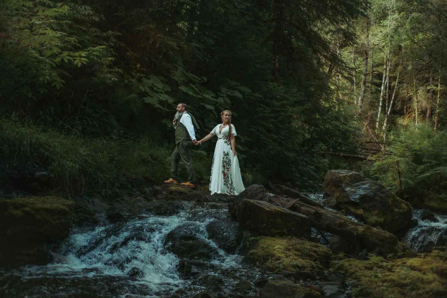 Two people in formal attire stand together on rocks beside a rushing forest stream in a moody woodland setting.