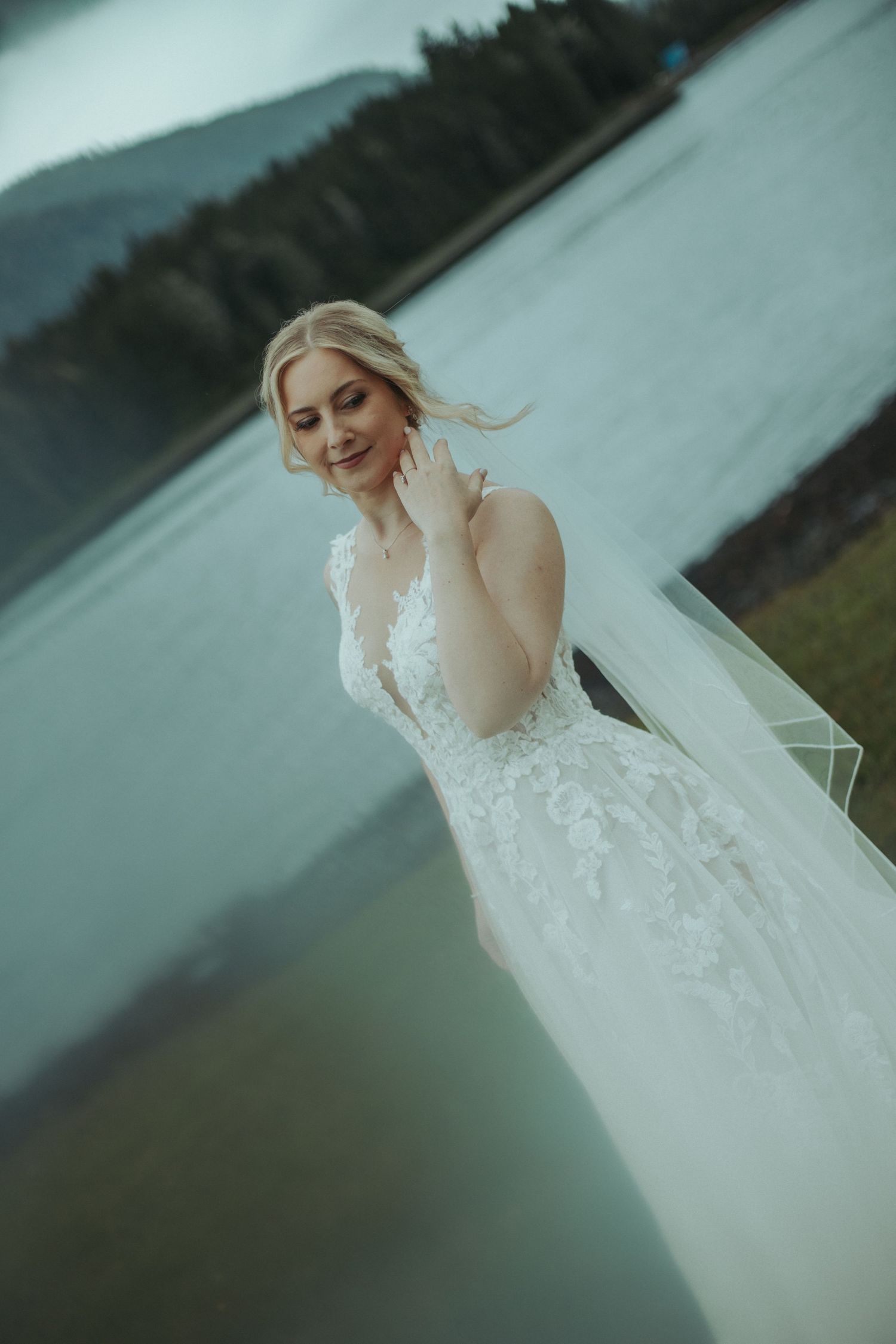 A bride in a white lace wedding dress stands by a lake on an overcast day in a series of artistic portraits.