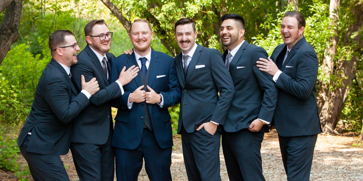 A group of groomsmen in navy suits share a candid laughing moment together in an outdoor garden setting.