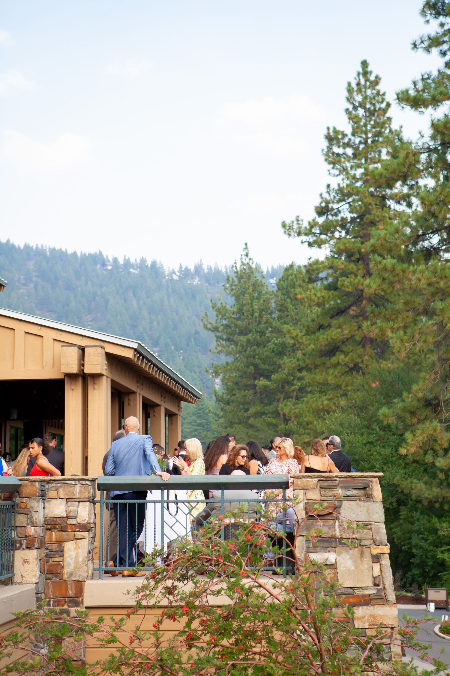 A group of people gather on a stone patio deck overlooking forested mountains at an outdoor venue.