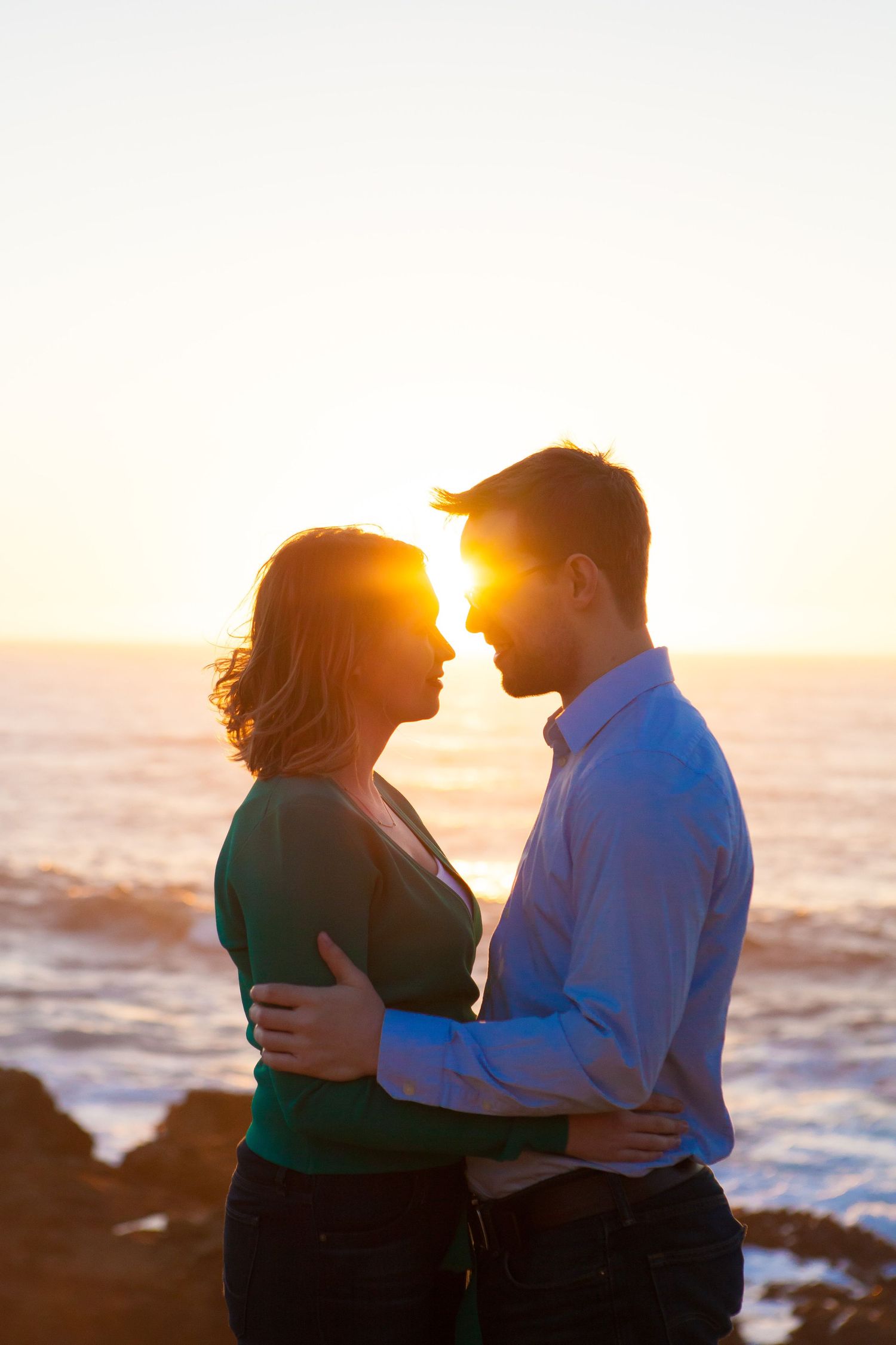 Silhouetted couple embracing on rocky beach at sunset with ocean waves in background.