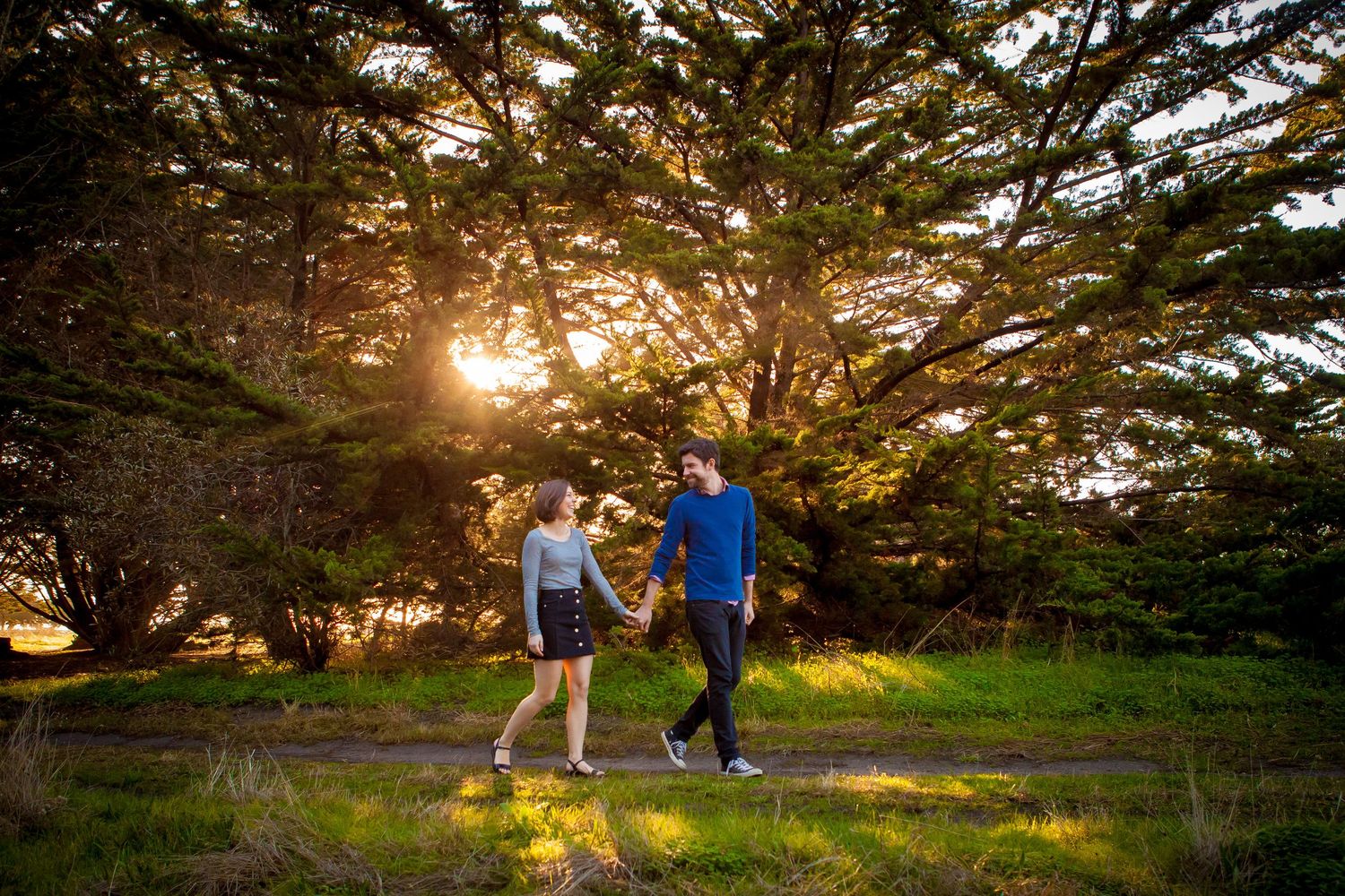 A couple walks hand in hand through a sunlit park with cypress trees during golden hour.