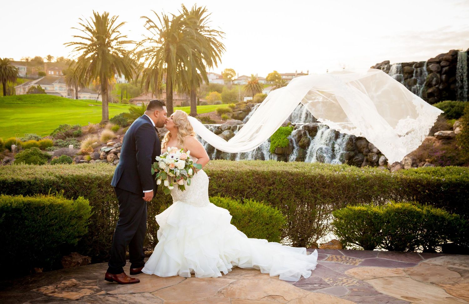 A flowing white wedding veil catches the wind at sunset against a backdrop of palm trees and manicured gardens.