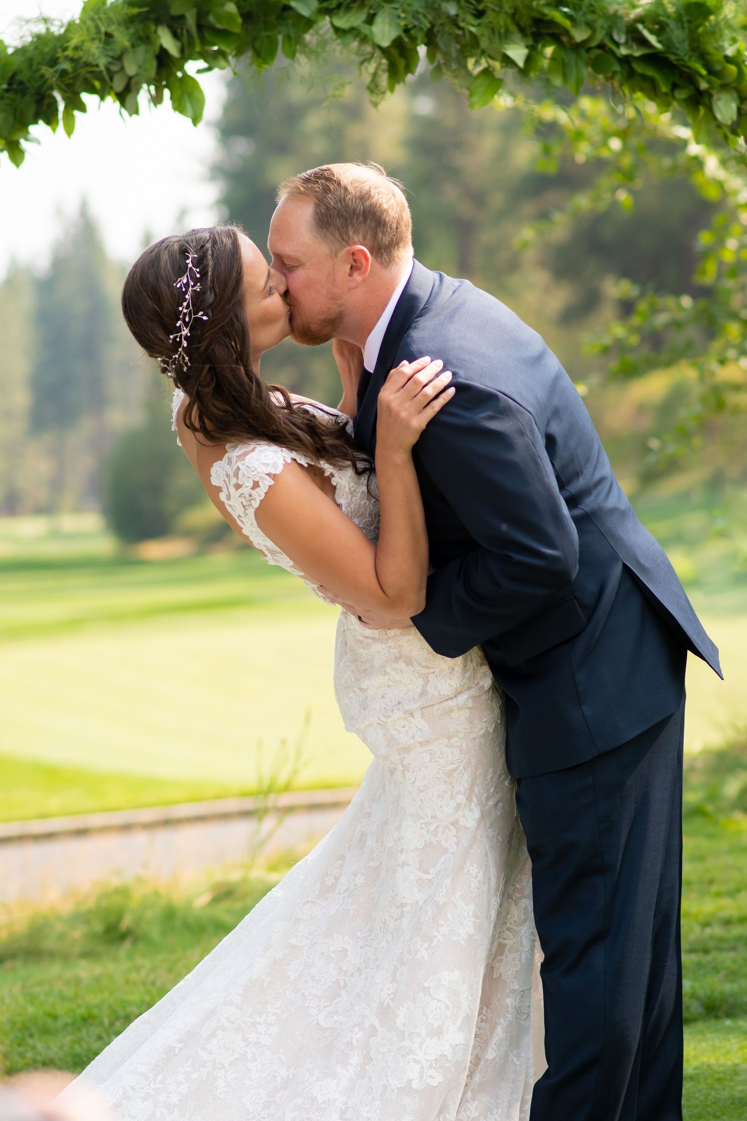 A romantic wedding couple share a kiss under green foliage in an outdoor garden setting.