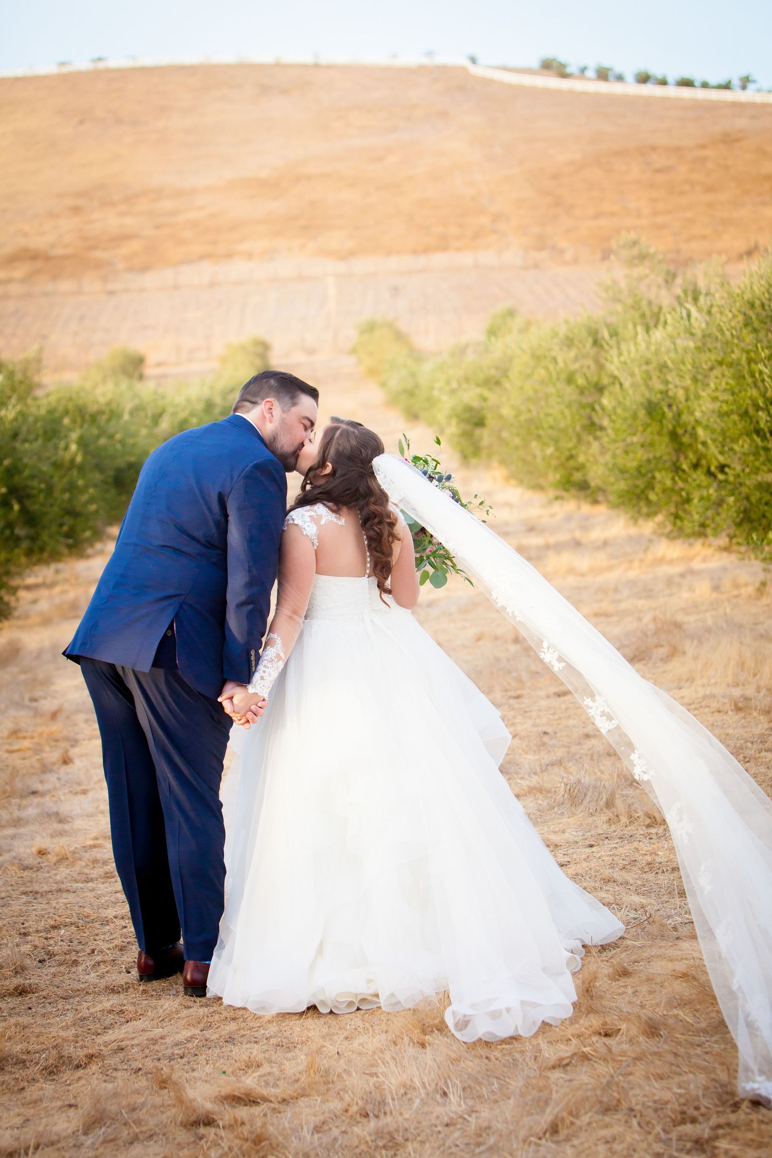 A romantic wedding couple shares a kiss on a golden hillside in matching navy and white attire.